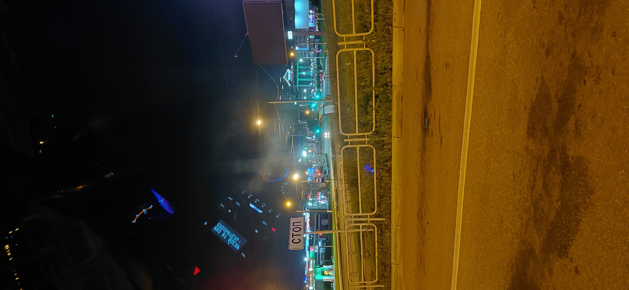 The image is a nighttime photograph taken from inside a vehicle, looking out at a street scene. A "СТОП" (STOP) sign is prominently displayed on the left side of the road. The scene appears to be a city street with various businesses and buildings illuminated by artificial lights. Traffic lights and streetlights are visible, along with the headlights of moving vehicles. The road surface is wet or reflective, suggesting recent rain or the presence of puddles. There is a yellow metal fence separating the road from a grassy area. In the distance, a large, illuminated building with a green facade stands out. The sky is dark, indicating it is nighttime, and there is some atmospheric haze or light pollution. The image is blurry in some areas, particularly in the upper portion, possibly due to the motion of the vehicle or camera settings. Without specific landmarks or identifying information within the image, it is not possible to definitively determine the location within Magnitogorsk, Russia. However, the presence of Cyrillic text on the "STOP" sign and other potential signage in the background suggests a Russian-speaking environment.