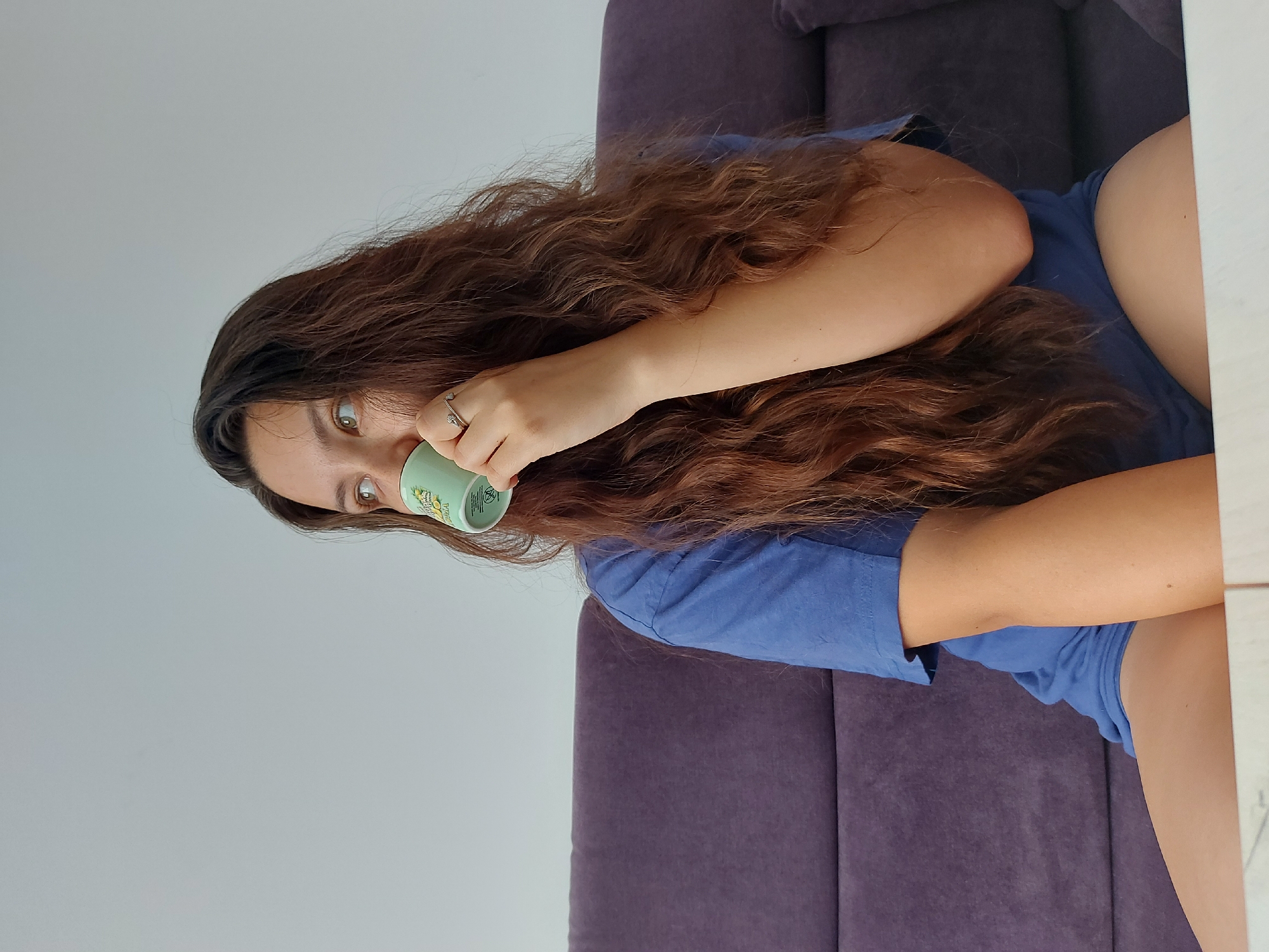 An indoor, portrait-style photograph, taken in Shumen, Bulgaria, features a young woman with long, wavy, reddish-brown hair. She is wearing a blue t-shirt and is seated on a dark purple sofa or armchair. The image is oriented such that the woman's head is tilted slightly to her right, and she looks towards the left side of the frame with a curious or sideways glance, her light-colored eyes prominent.
She holds a light green mug with both hands, raising it to her mouth as if drinking. The mug has a decorative pattern of yellow and green shapes, possibly citrus fruits like lemons or limes, and some partially visible, unreadable text. A silver ring with a decorative element is visible on the ring finger of her right hand.
The background to her left consists of a plain, light grey wall, while a white surface, possibly a table or furniture, is visible on the right edge of the frame. The lighting is soft and even, suggesting daytime indoors. The overall mood is relaxed and casual.
