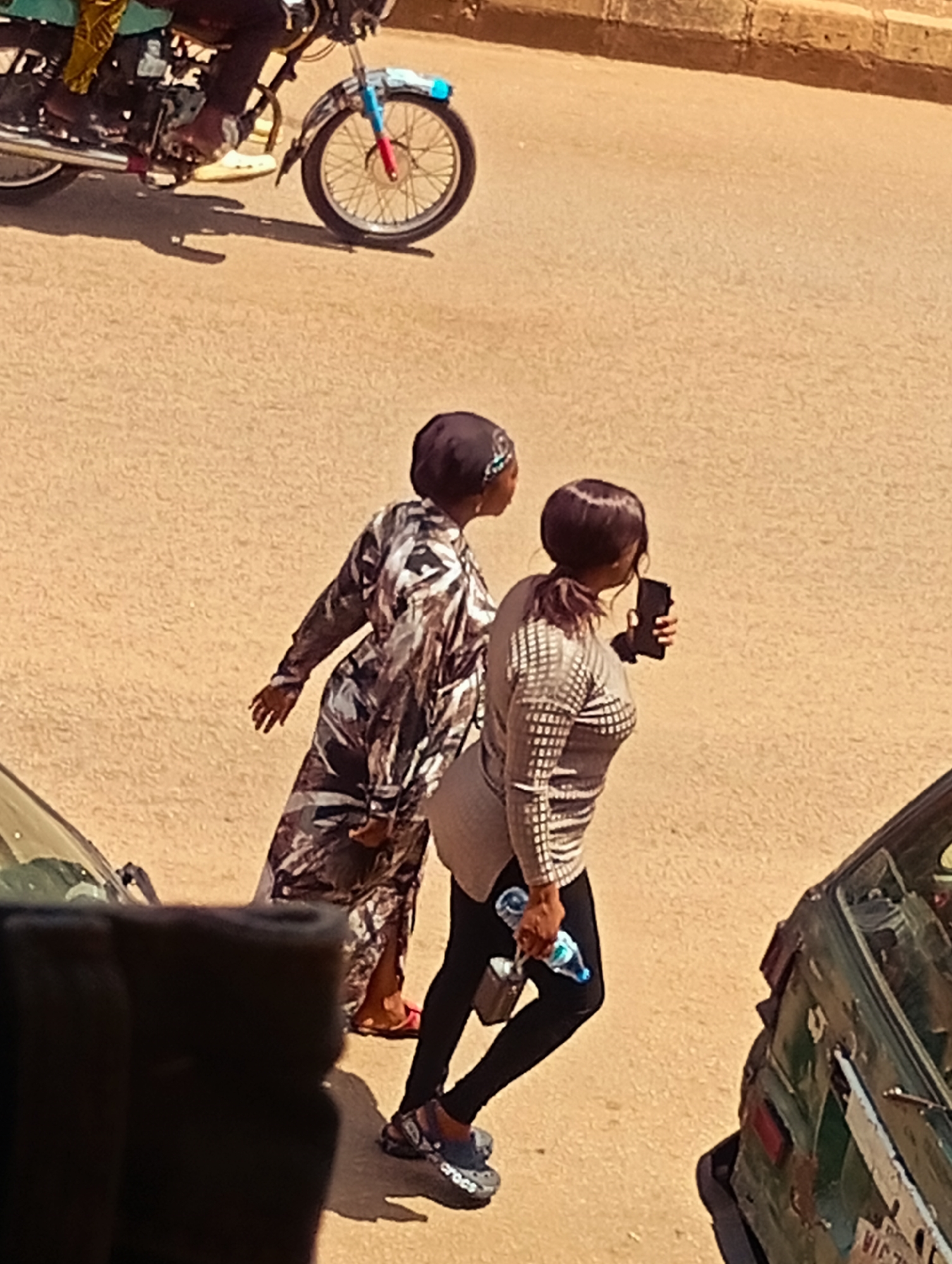 This is an outdoor, eye-level, medium shot of a street scene in Bukuru, Nigeria. The sun is high in the sky, casting distinct shadows, suggesting it's midday. The ground is a dusty, tan-colored asphalt.
In the foreground, the top of a dark vehicle is visible, out of focus.
The main subjects are two women walking. The woman on the left wears a dark headscarf and a long, flowing dress with a black, white, and grey marbled pattern. Her feet are adorned with bright red sandals. She is walking with her left hand extended slightly behind her.
The woman on the right wears a dark headscarf and a fitted, long-sleeved shirt with a checkerboard-like pattern in shades of grey. She wears black leggings and dark grey Crocs-style shoes. She is holding a dark smartphone in her right hand, appearing to take a photo or video, and a clear plastic water bottle with a blue label in her left hand.
In the background, the rear portion of a motorcycle is visible. It has a chrome exhaust pipe, a spoked wheel with a partially blue and red rim, and a fender adorned with blue and red details. A person's leg, wearing dark trousers and light-colored shoes, is visible on the motorcycle.
To the right of the women, the back of a green car is visible, appearing weathered and possibly damaged, with a license plate that has some visible text, including "VIC" and possibly numbers.
The overall scene depicts everyday street activity in an urban environment.