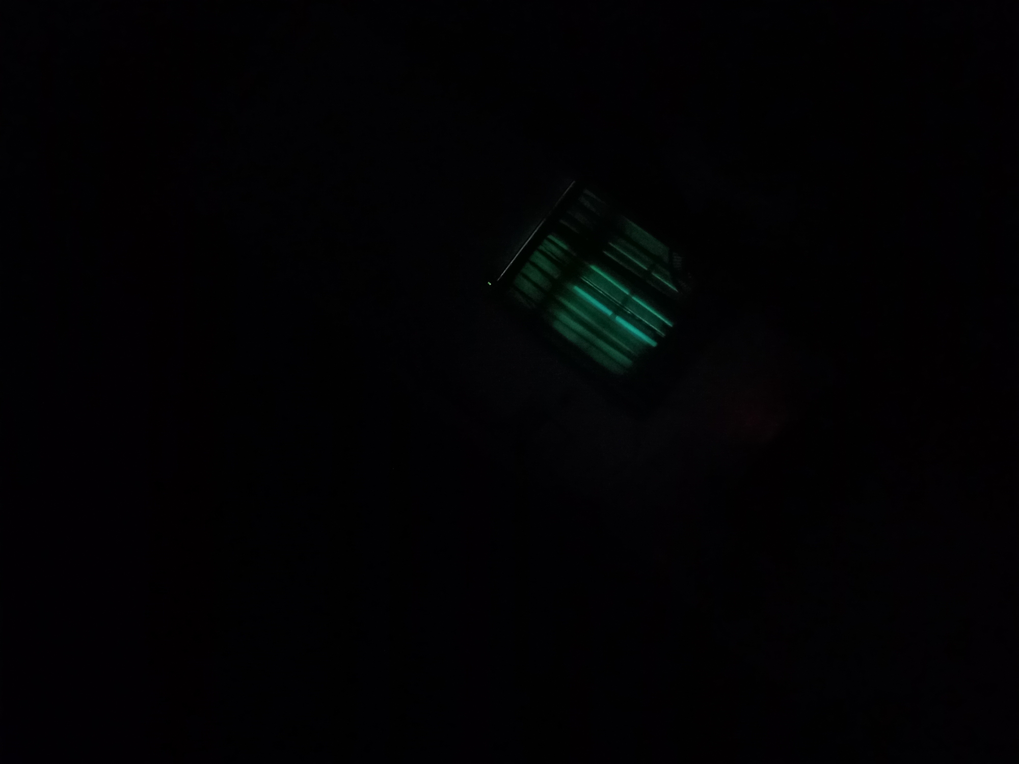 A close-up, high-angle shot shows a cylindrical object with a ribbed texture emitting a green light in a dark environment. The object is angled and positioned in the lower right quadrant of the frame, with the majority of the image being black. No other details are visible in the scene. The location in the city of Pajagalan, Indonesia, is not discernible from the image.