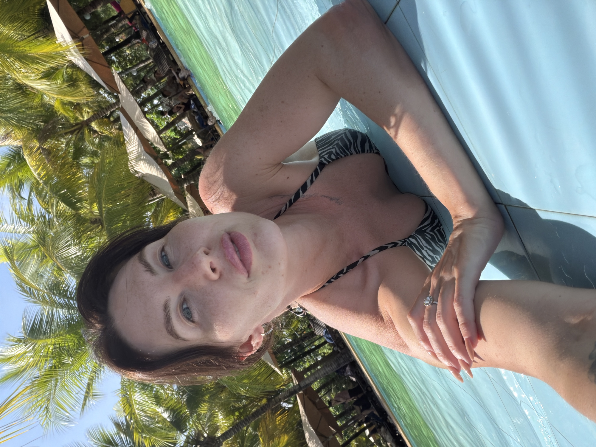 A woman with brown hair and fair skin is lying in a swimming pool. She is wearing a black and white zebra-print bikini. Her left arm is resting on the edge of the pool, and her right arm is bent at the elbow, with her hand on her chest. She is wearing a diamond ring on her left ring finger. The pool water is turquoise. In the background, there are palm trees, lounge chairs, and umbrellas, suggesting a tropical resort setting. The sky is blue. The overall scene suggests a leisurely day at a resort.