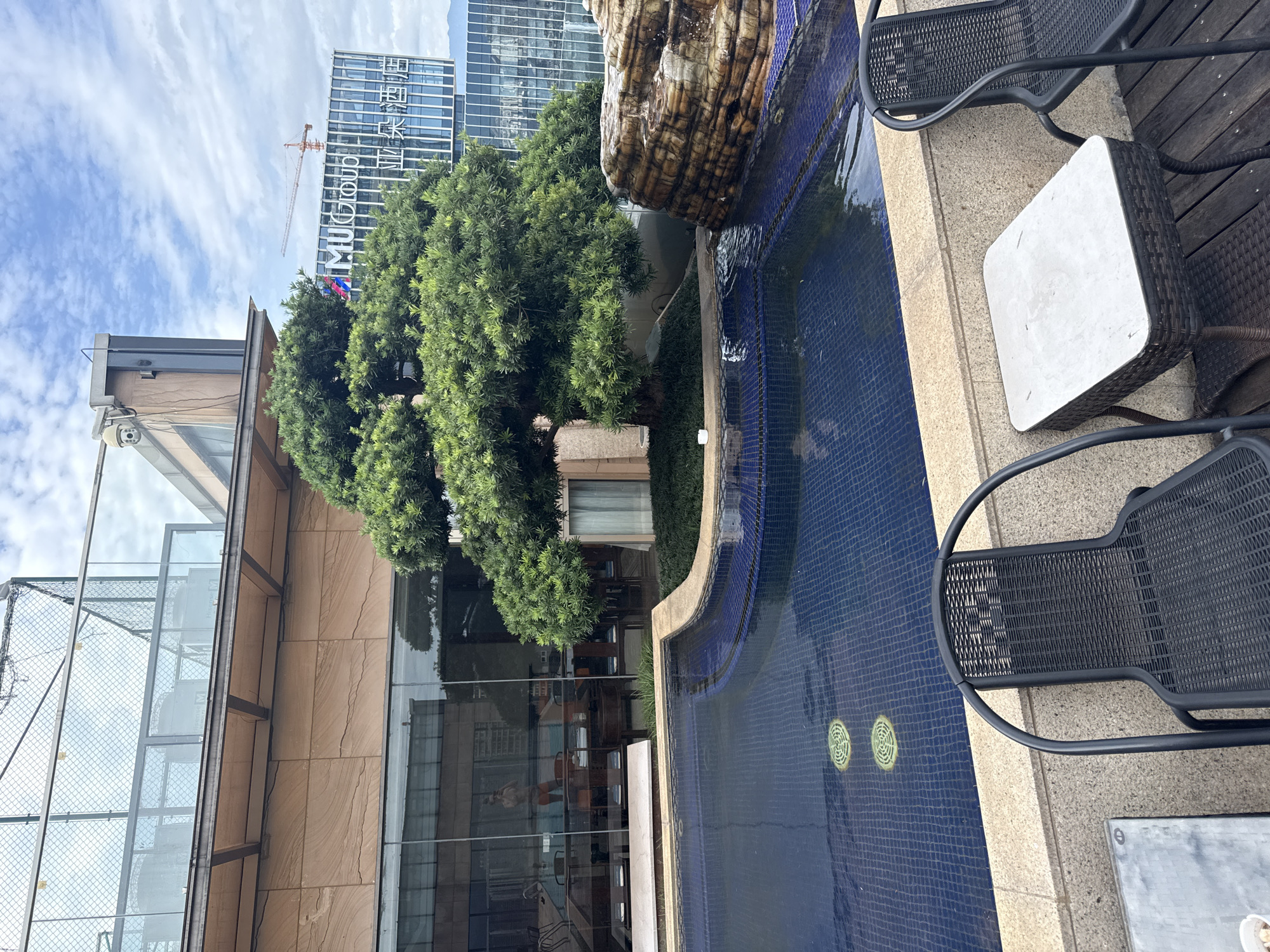 This is a photograph taken from a rooftop setting in Yiwu, China. In the foreground, a dark blue tiled swimming pool occupies the majority of the lower portion of the image. Two black outdoor chairs with woven seats and backs are positioned at the edge of the pool on a wooden deck. A small, light-colored square table with a dark woven base sits between the chairs.
Behind the pool, a large, lush green tree with dense foliage is situated in the center of the frame. To the right of the tree, a rock formation with layered textures and a small waterfall feature is visible, with water flowing into the pool.
In the mid-ground, a building with a glass facade reflects the surrounding environment, including what appears to be another pool area and seating arrangements indoors. Further in the background, a modern building with the prominent "M U Group" logo and Chinese characters is visible, with a construction crane also present. The sky above is partly cloudy, with a mix of blue sky and white clouds. The overall scene suggests a hotel or resort rooftop amenity in an urban environment.