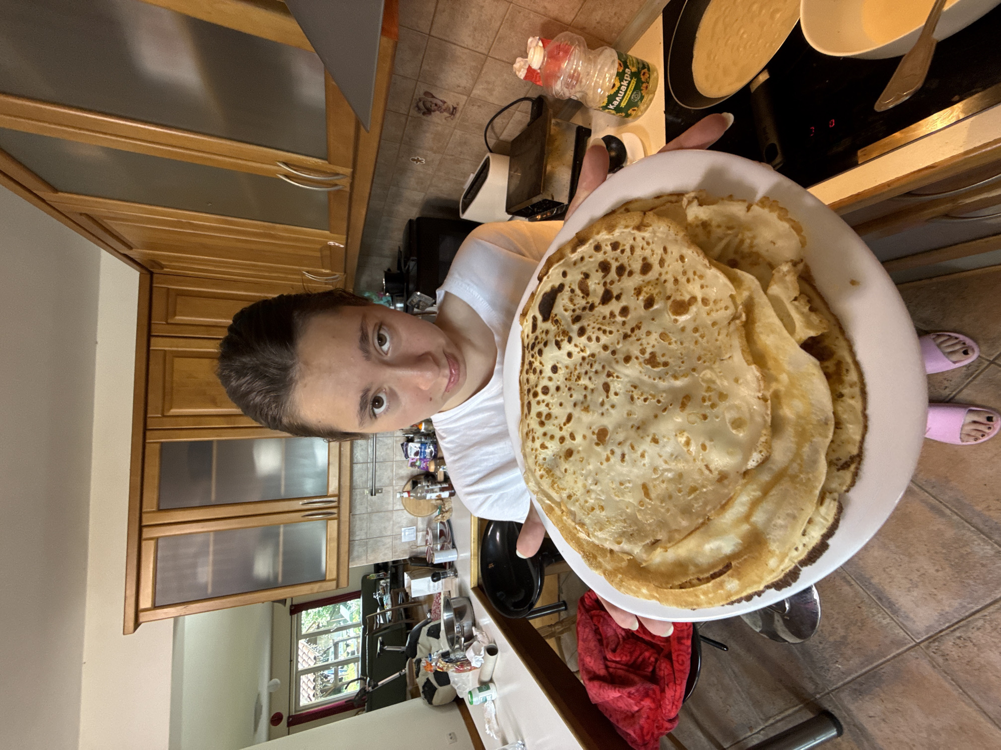 A young woman with dark hair pulled back, wearing a white t-shirt and pink slippers, stands in a kitchen holding a large white plate piled high with crepes. Her expression is neutral, and she is looking directly at the camera. The kitchen features wooden cabinets with glass-fronted doors, a tiled backsplash, and a countertop with various items including a bottle of cooking oil labeled "Калиакрa" (KALIACRA), a toaster, and a frying pan with batter. The floor is tiled in a beige and brown pattern. The background shows a window with a view of greenery and possibly other buildings, suggesting an urban or suburban setting, consistent with a residential kitchen in Sofia, Bulgaria.