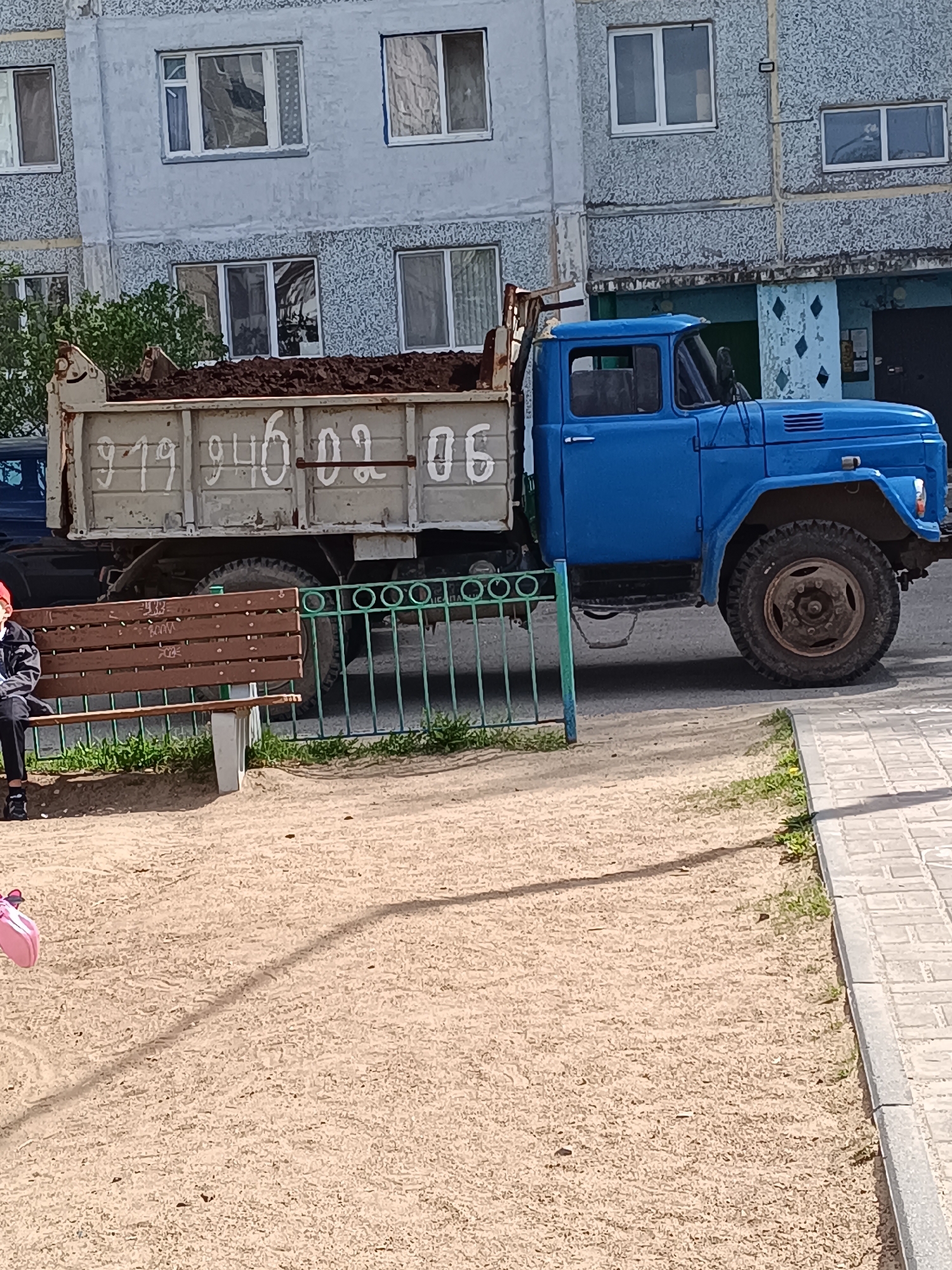 A photograph taken in Tobolsk, Russia, depicts a blue ZIL-130 truck with a weathered, gray dump bed filled with dark material, likely soil or construction debris. The truck bears the inscription "919 940 02 06" in white on its bed. Adjacent to the truck, a brown wooden bench sits beside a green metal fence, with a person in dark clothing seated on the bench. A pink bag rests on the ground near the bench. The foreground shows a sand surface with sparse vegetation. The backdrop features a multi-story, gray apartment building with numerous windows. The building exhibits a weathered facade, and one entryway is visible with a distinct teal-colored overhang and teal diamond-shaped decorations. The overall impression is of an urban, residential area showing signs of age and wear.