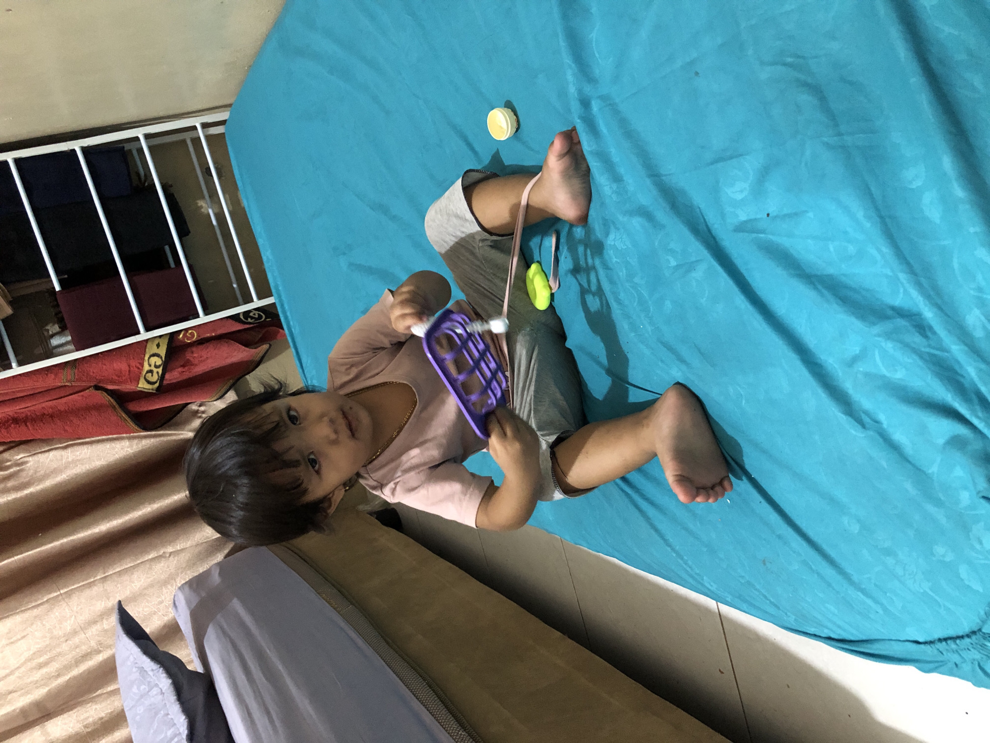 A young girl sits on a teal-colored mattress with a purple toy in her hands. She is wearing a light pink t-shirt and grey pants. To her left, a portion of a bed with a grey pillow is visible. In the background, there are beige curtains, a red towel, and a white rack holding folded fabrics. The environment appears to be indoors, possibly a bedroom. The image was taken in Pashiebeutong, Indonesia.