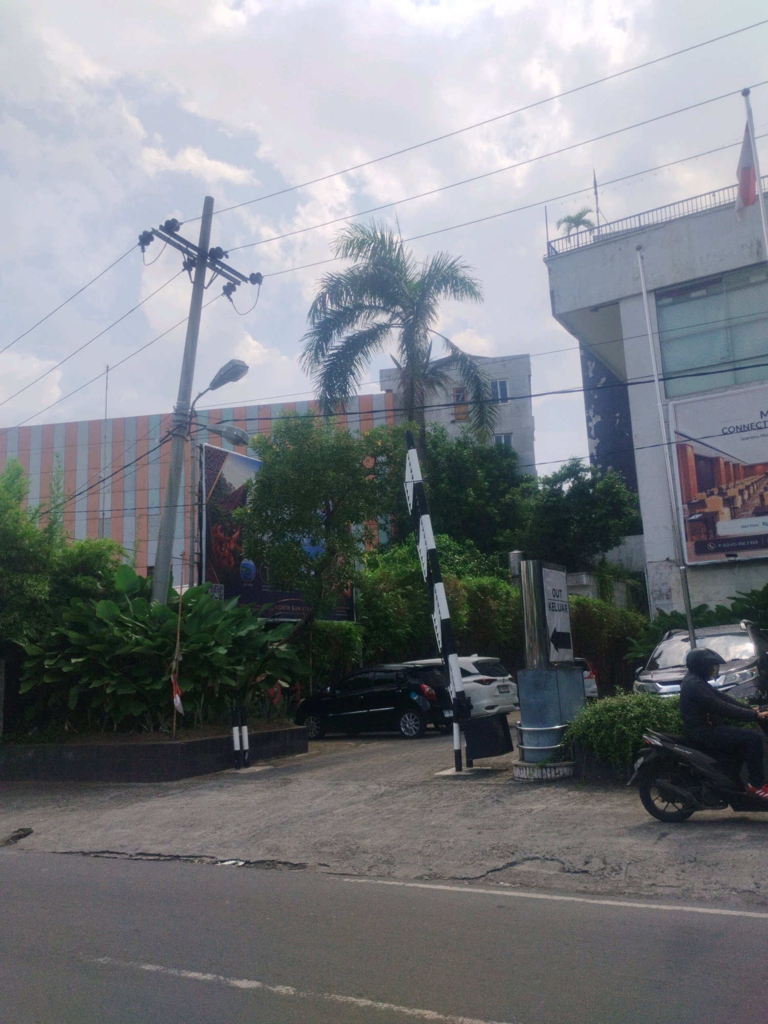 The image depicts an outdoor scene in the city of Medan, Indonesia, on a cloudy day. In the foreground, a gray asphalt road with visible cracks is present. Beyond the road, a paved area leads to a building entrance. Several vehicles are parked or in motion. A black motorcycle with a rider wearing a black jacket, helmet, and red-accented shoes is visible on the right, moving towards the right. A white SUV is parked behind a striped barrier. A black car is also parked in the background. A metal barrier with black and white stripes is positioned to control entry and exit. A sign with text, including "OUT KELUAR" and an arrow pointing to the right, is mounted on a pole.
Several buildings are visible in the background. On the right, a multi-story building with a white facade and dark accents features a balcony with a small palm tree and a flag on top. A large billboard advertising "CONNECTORS" is attached to this building. To the left of this, a building with vertical striped cladding in shades of orange, pink, and white is visible. Further to the left, a tall, gray building with multiple windows stands under a cloudy sky.
Abundant green foliage, including large-leafed plants and trees, surrounds the entrance area and borders the paved surfaces. A utility pole with multiple wires and streetlights stands prominently on the left side of the image, with one streetlight appearing to be a modern LED design. The overall scene suggests a commercial or mixed-use area in Medan.