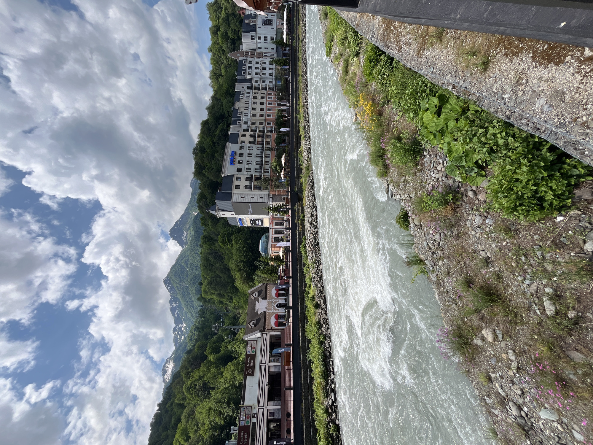 The image captures a scenic view of a fast-flowing river in Estosadok, Russia. The river, characterized by its turbulent, whitewater rapids, occupies the foreground and middle ground of the frame. Its banks are lined with a mix of rocks, gravel, and lush green vegetation, including small flowering plants.
In the background, a row of buildings lines the opposite bank of the river. These structures appear to be hotels or resorts, with architectural styles that suggest a blend of modern and traditional elements. One prominent building features multiple stories with balconies and a distinctive dark-tiled roof. To the left of these buildings, a mountain range rises, covered in dense green forest, with snow-capped peaks visible in the distance. A cable car system with multiple cabins can be seen ascending the mountainside.
The sky is partly cloudy, with large white cumulus clouds against a blue backdrop, indicating fair weather. The overall impression is one of a vibrant natural landscape complemented by human development in a mountainous resort town.