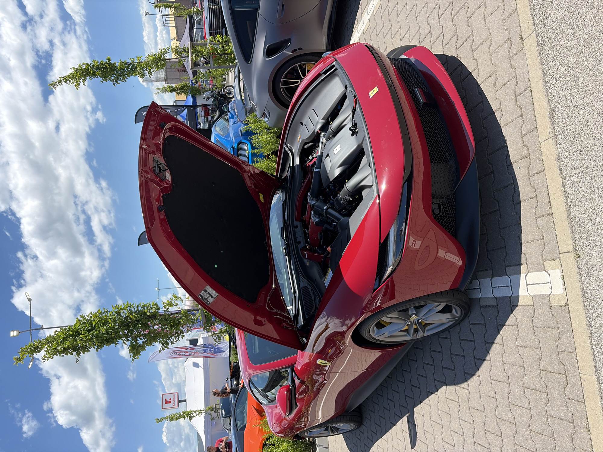 A bright red Ferrari Roma is parked in an outdoor lot, its hood open, revealing its engine. The car is positioned at a slight angle, with its front facing the viewer's right. The car's glossy finish reflects the bright sunlight and the surrounding environment. In the background, other vehicles are visible, including a dark grey Porsche 911, a blue Ford Mustang, and an orange sports car. Several people are present, some standing near the cars and others walking in the background. Tents and banners are set up, suggesting an outdoor event or exhibition. Tall, slender trees with lush green foliage line the perimeter of the lot, and a bright blue sky with scattered white clouds forms the backdrop. The ground is paved with interlocking grey bricks, with white parking lines marking the spaces. The scene is illuminated by natural daylight, casting distinct shadows on the paved surface. The overall atmosphere is that of a car show or automotive gathering, likely held in an open area with amenities.