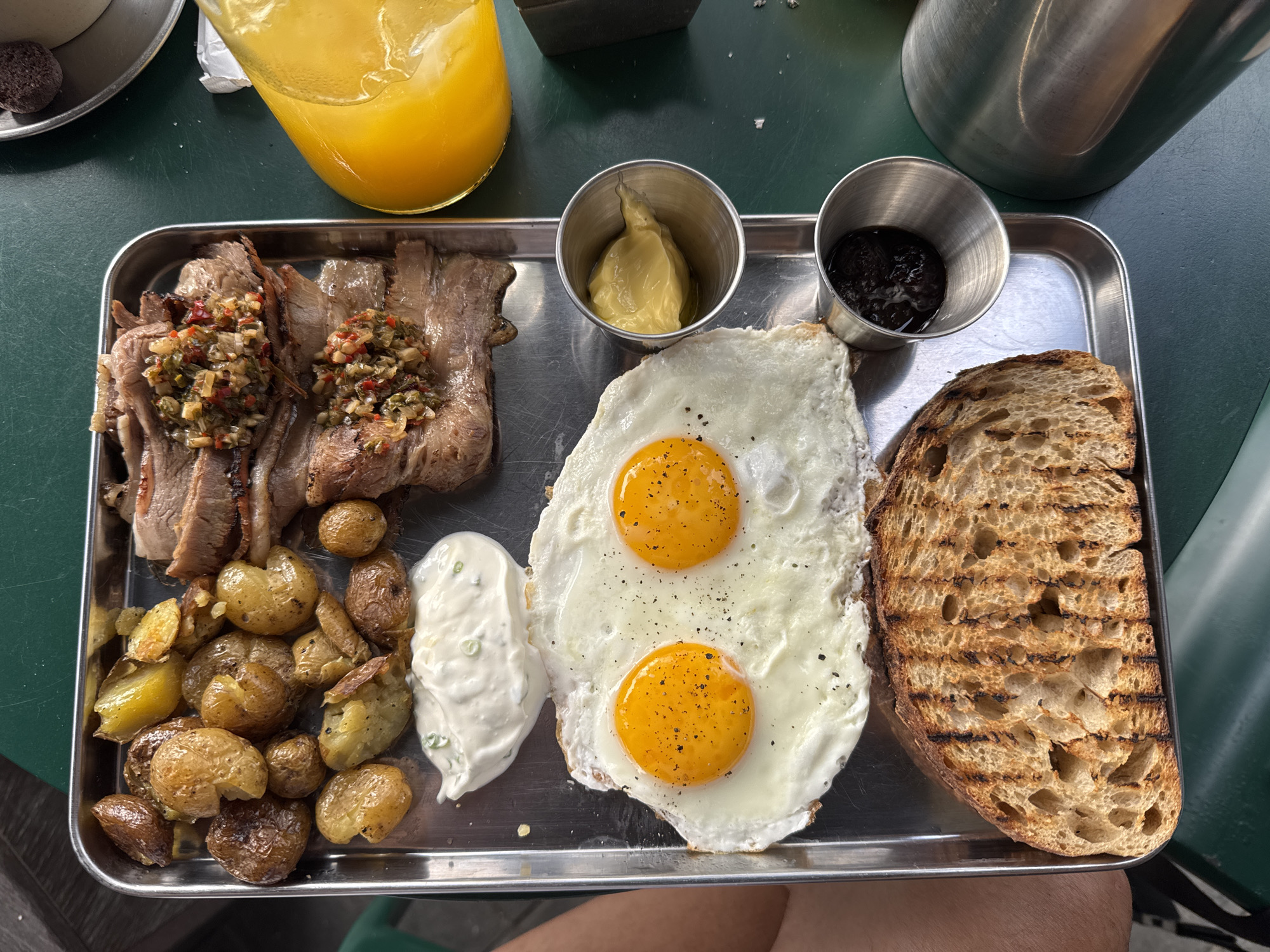 The image shows a metal tray containing a meal, likely taken in North Kuta, Indonesia. The meal consists of sliced beef with a colorful topping, roasted potatoes, two fried eggs, a dollop of white sauce with green flecks, a pat of yellow butter, a dark-colored jelly or jam, and a slice of toasted bread. A glass of orange juice is visible in the upper left corner, along with a cup of coffee with a chocolate cookie on the side, and a metal thermos is visible in the upper right corner of the image. The tray is sitting on a green table. A small portion of a person's legs is visible at the bottom of the image.