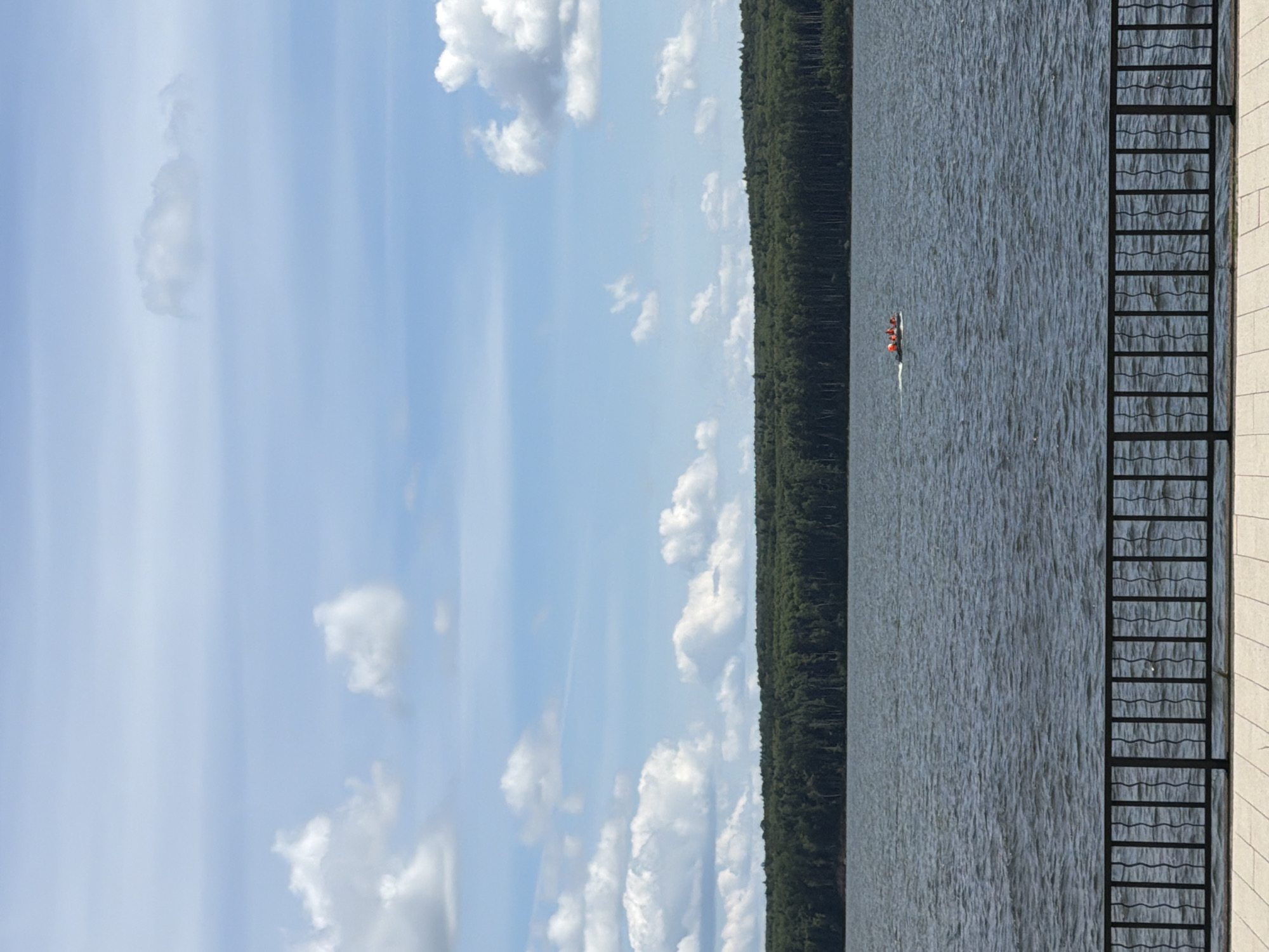 The image depicts a wide shot of the Kama River in Chaikovsky, Russia, on a bright, cloudy day. In the foreground, a black metal railing with a wavy design stands atop a beige tiled walkway. The river occupies the middle ground, its surface rippled by a gentle breeze. In the distance, a dense forest of green trees stretches along the horizon, meeting the blue sky filled with scattered white clouds. A small inflatable boat with several people wearing orange life jackets is visible on the river, moving from left to right. The overall scene conveys a peaceful, natural setting.