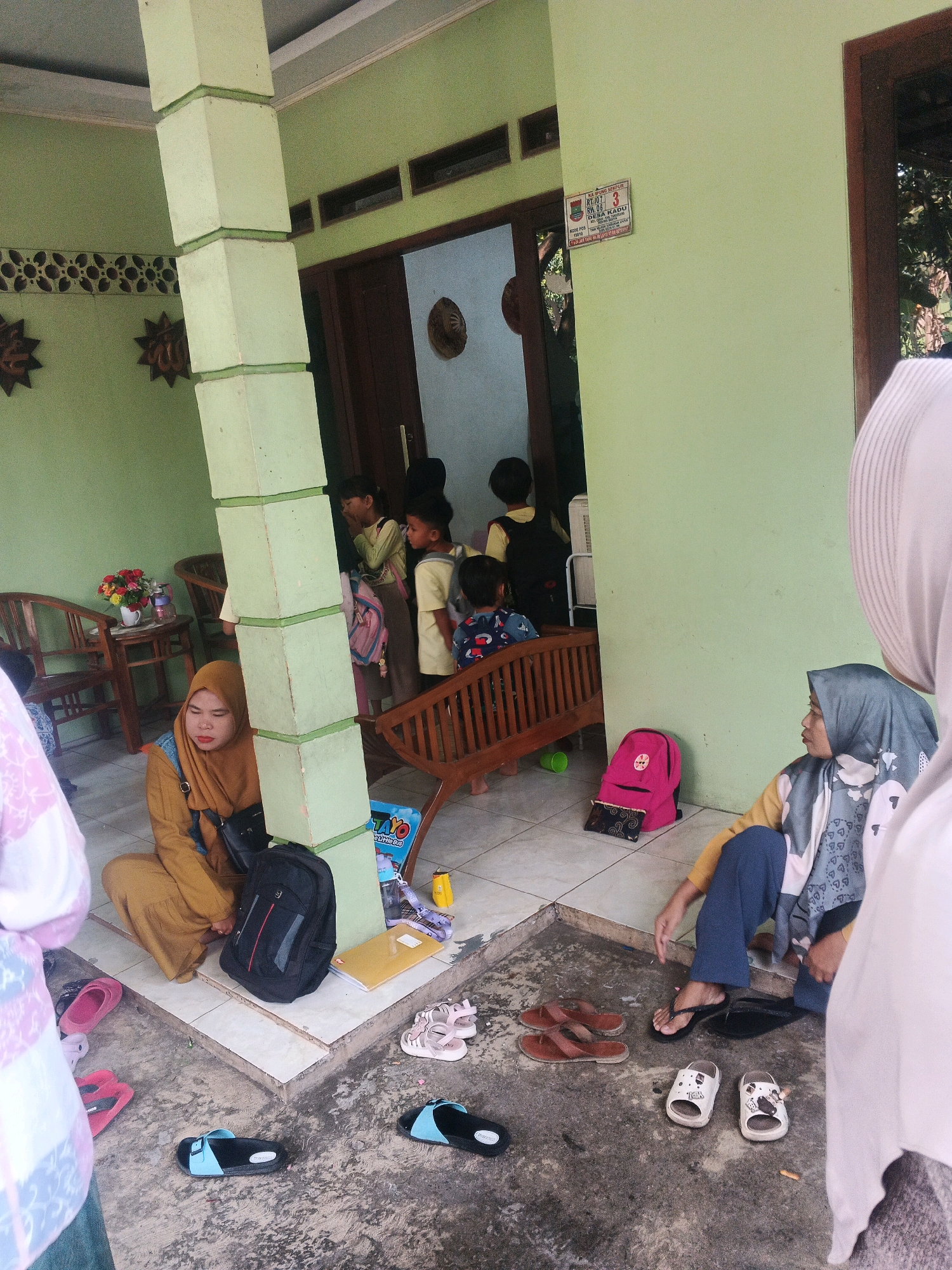The image depicts an outdoor scene in the city of Kadu, Indonesia, specifically in front of a house with a light green exterior wall. A large, segmented light green concrete pillar stands prominently in the foreground, supporting an overhang. To the left, there is a wooden chair and a small table with a vase of flowers.
Several people are present. In the center, a woman wearing a mustard-colored hijab and a denim vest sits on the ground. A black backpack rests beside her. To her right, another woman, also wearing a hijab (this one grey and white patterned), is seated on the ground and looking to her left. A pink backpack is visible near her feet.
In the background, several children are gathered near an open doorway. Some are wearing yellow shirts and dark pants or skirts, and appear to be carrying backpacks. One child is seated on a wooden bench. The doorway leads into a darker interior space.
The ground is a combination of tiled flooring and concrete. Various footwear, including sandals and flip-flops, are scattered on the ground around the seated individuals. A yellow folder and a children's book titled "Tayo" are also visible on the ground. The overall impression is a casual gathering or waiting area outside a residential building in Kadu.