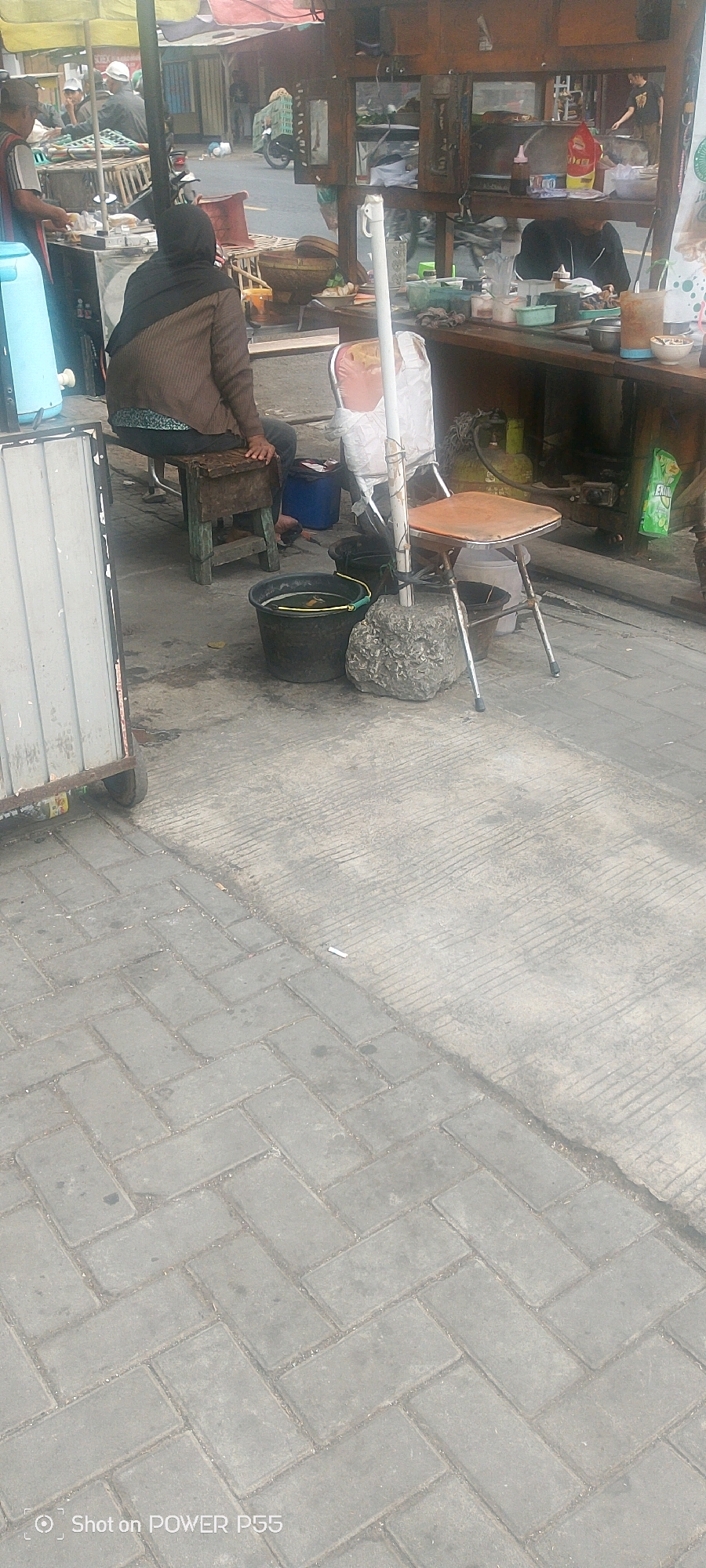 In an outdoor urban setting, a woman wearing a dark headscarf and a brown jacket sits on a wooden stool. She is positioned to the left of a folding chair with a light-colored cushion, which is set back from a wooden food stall. In front of the woman, a dark bucket with a yellow handle is visible, containing water and possibly some items. To the right of the folding chair, another, smaller bucket is present. The food stall displays various items, including bottles of sauces and what appears to be food in containers. A large gray rock anchors the base of a pole supporting a white fabric, situated near the folding chair. The ground is paved with interlocking grey rectangular blocks, with a section of concrete to the right marked with parallel lines. A cigarette butt is present on the concrete surface. In the background, the food stall structure extends upwards, revealing shelves with items, and a glimpse of a motorcycle and other people is visible beyond the stall.