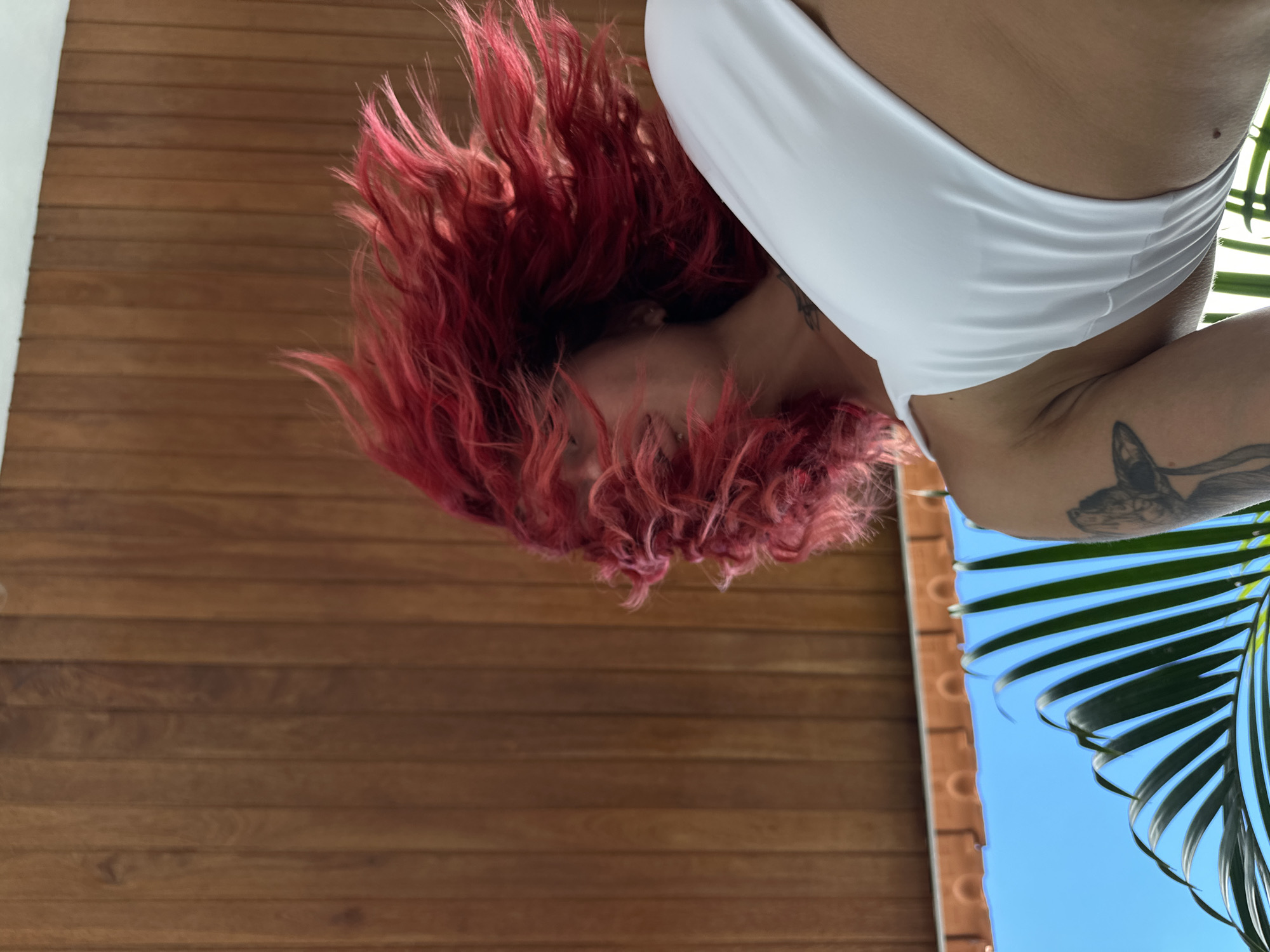 The scene is a low-angle shot looking upwards at a woman with red, curly hair. She's wearing a white bikini top. A tattoo is visible on her upper arm. The background consists of horizontal wooden planks and a palm frond against a bright blue sky. Given the prompt's mention of North Kuta, Indonesia, it is reasonable to infer the location is there. The overall impression is a casual outdoor setting, possibly a poolside or tropical resort.
