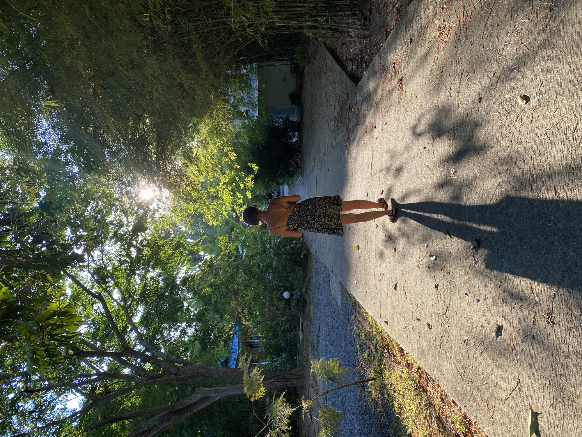 A young woman wearing a floral dress walks away from the camera on a concrete pathway surrounded by lush greenery. Sunlight filters through the leaves of trees and bamboo, casting long shadows across the path. In the background, a small building with a blue roof is visible, along with bicycles. The scene appears to be a serene, tropical environment in ตำบลเกาะพะงัน, Thailand.