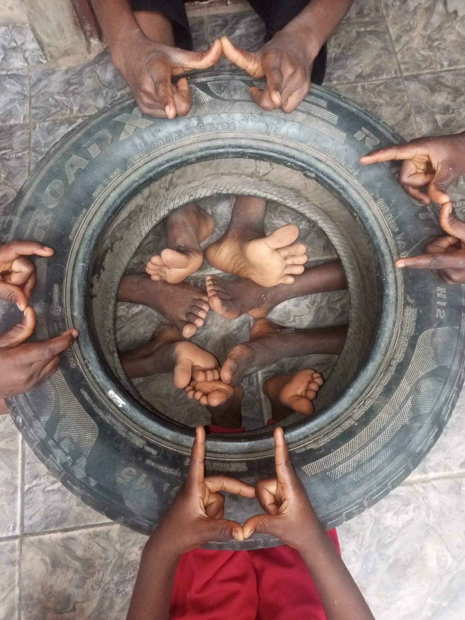The image shows an overhead view of a used tire on a tiled floor. Inside the tire are several pairs of bare feet. Four pairs of hands surround the tire, each making a finger gesture of unknown meaning. The tire has "ROADX" printed on it, along with other tire specifications, including "MADE IN CHINA." The letters "H12" and the marking "DOT KEAP FJJ 37" are visible on the tire as well. The location of the image is not specified and therefore not confirmed. There is no reference to Tempure, Nigeria, in the image.