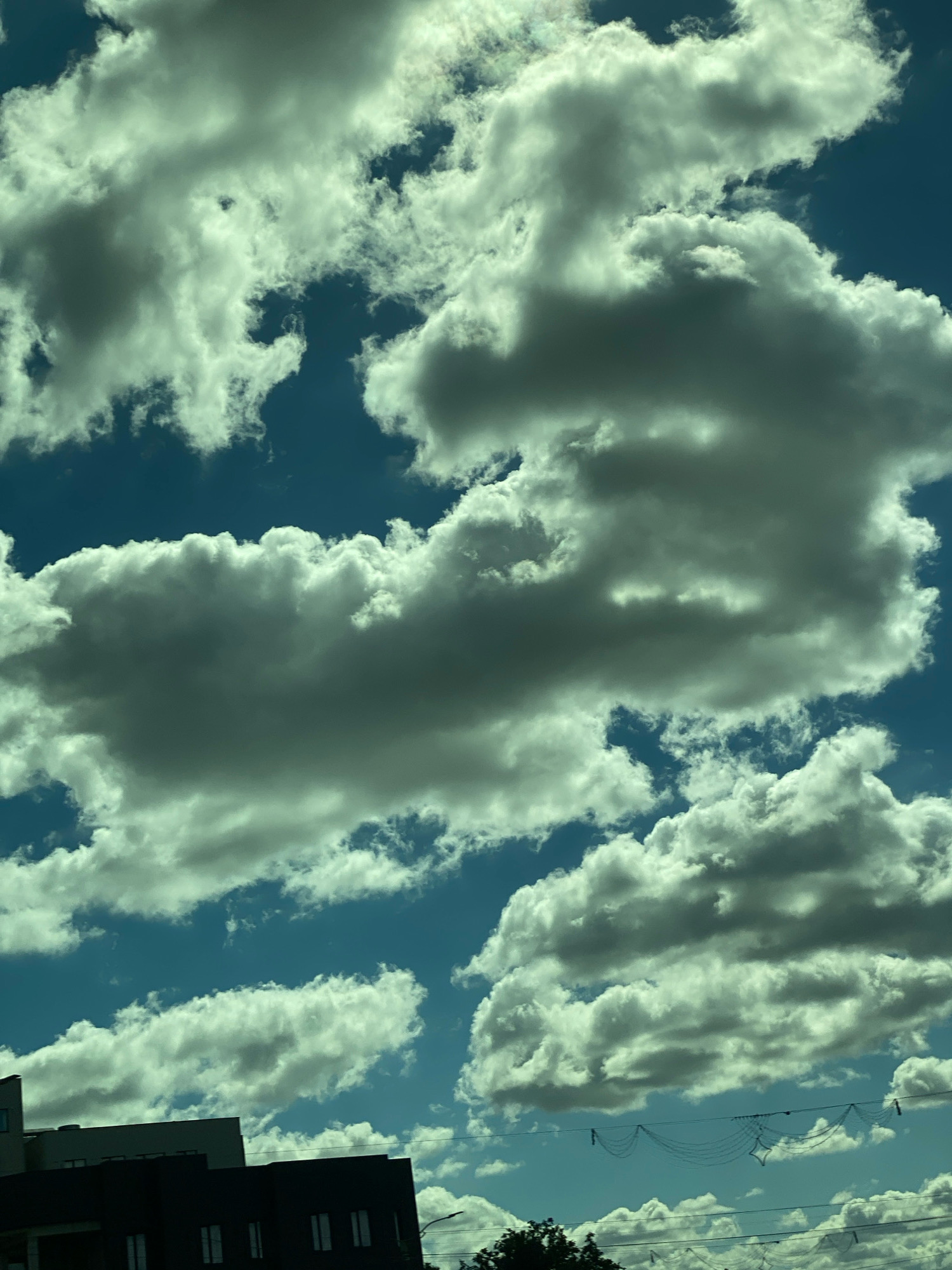 The image shows a daytime sky with dramatic, voluminous cumulus clouds. The sky is a deep blue, and the clouds are a mix of bright white where illuminated by the sun and darker grey in their shadowed areas. In the lower left quadrant of the image, the tops of two buildings are visible. The building in the foreground is dark, with multiple windows visible. The building behind it is lighter in color. In the far distance, a string of decorative lights or netting is visible spanning across the frame, with additional utility wires also present. A dark mass of tree foliage is present in the lower right corner. The scene appears to be an outdoor urban environment, possibly a city like Chișinău, Moldova, based on the architectural style of the buildings and the presence of overhead wires.