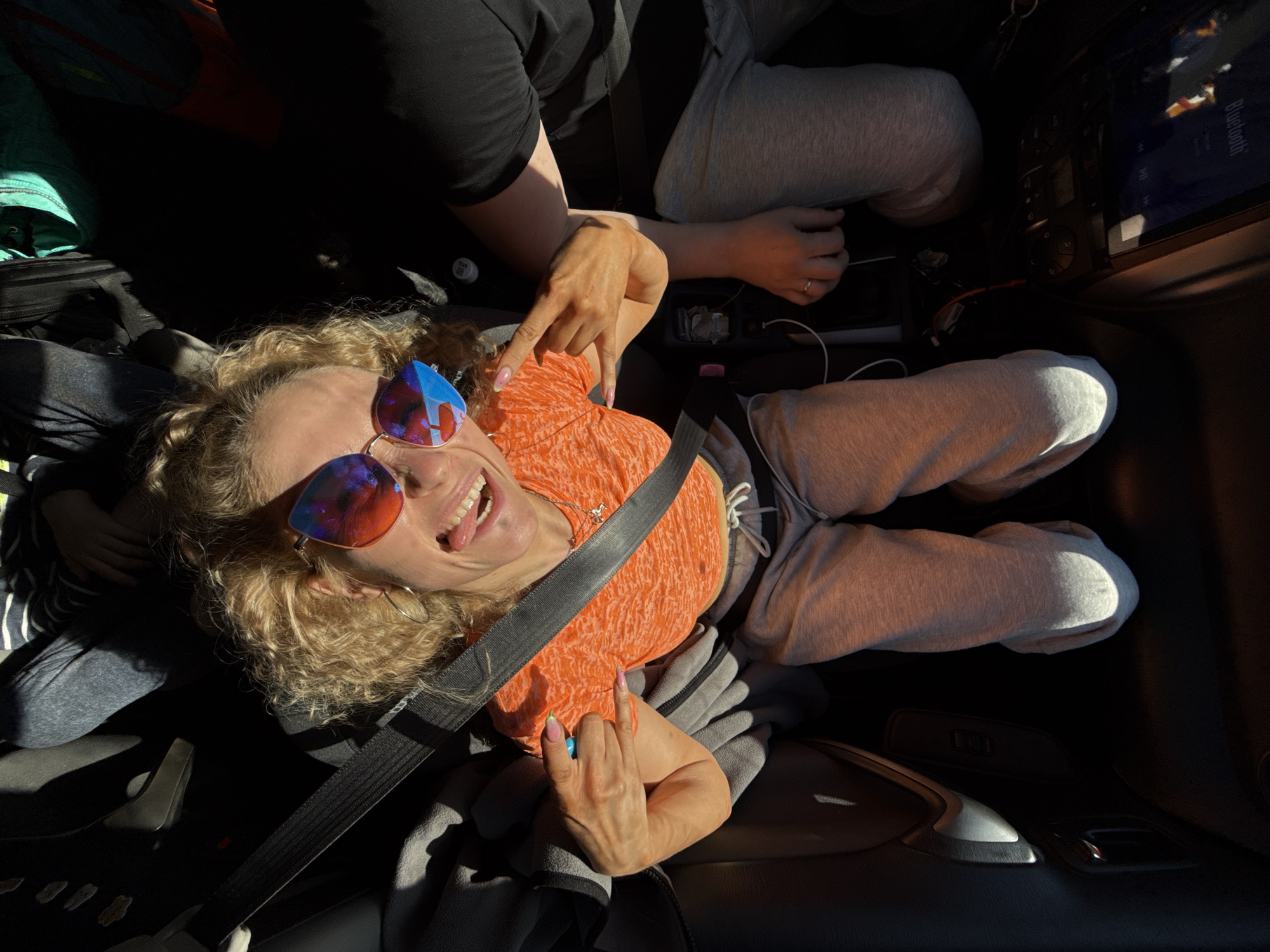 The image shows a woman inside a car, wearing an orange shirt, grey sweatpants, and sunglasses. She is wearing a seatbelt. Another person is visible in the image, also wearing sweatpants, and a black shirt, likely in the passenger seat. The car's infotainment system is on. The location is possibly Chaikovsky, Russia, but this is not visually confirmed.