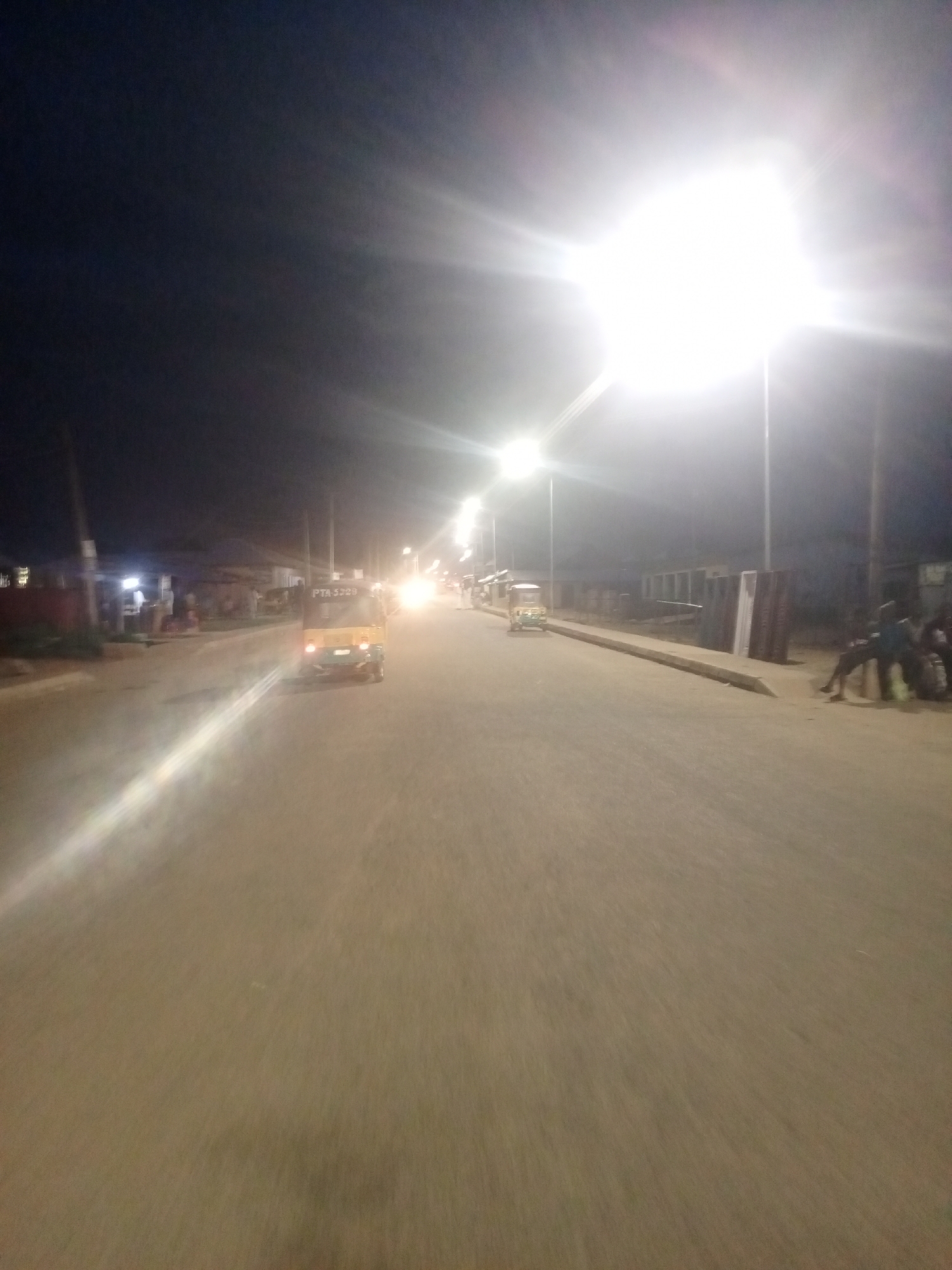 A motion-blurred, night-time photograph taken on a street in Bukuru, Nigeria, shows a paved road with streetlights illuminating the scene. A yellow and green auto-rickshaw, identified by the license plate "PTA 5233", is in the foreground, moving away from the camera. Several other vehicles and streetlights are visible further down the road, suggesting an urban environment. On the right side of the road, a group of people are seated, and on the left, buildings and what appear to be construction materials are present. The bright glare of a streetlight dominates the upper right portion of the image.