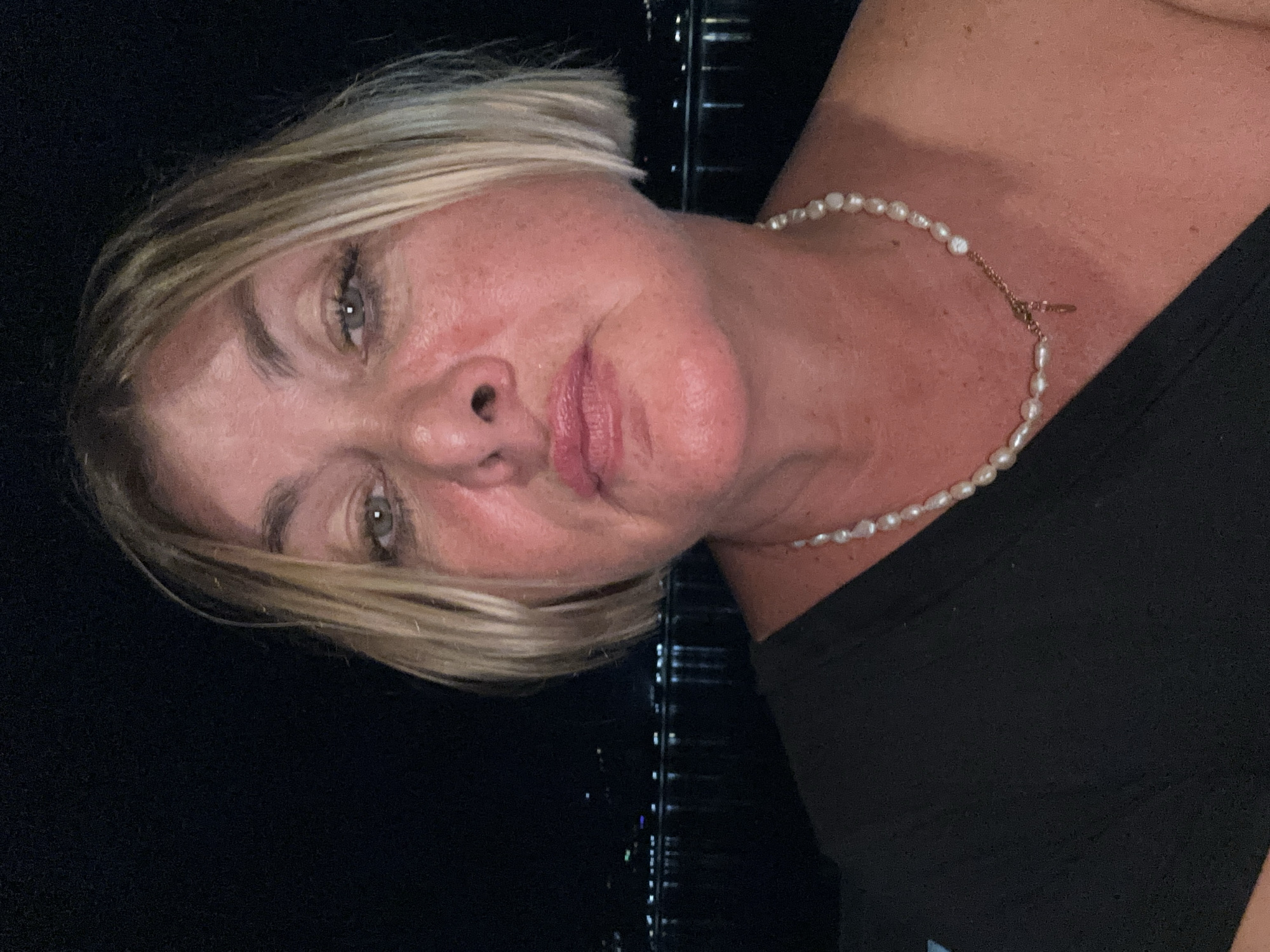 A portrait of a woman with short blonde hair wearing a pearl necklace is set against a dark, indistinct background. The woman has fair skin, grey eyes, and a neutral expression. The dark background suggests an outdoor setting, likely at night. Vertical lines are visible in the background, possibly indicative of a structure. The scene appears to be in บ้านบางรักษ์, Thailand, though there is no specific identifiable feature to confirm this.