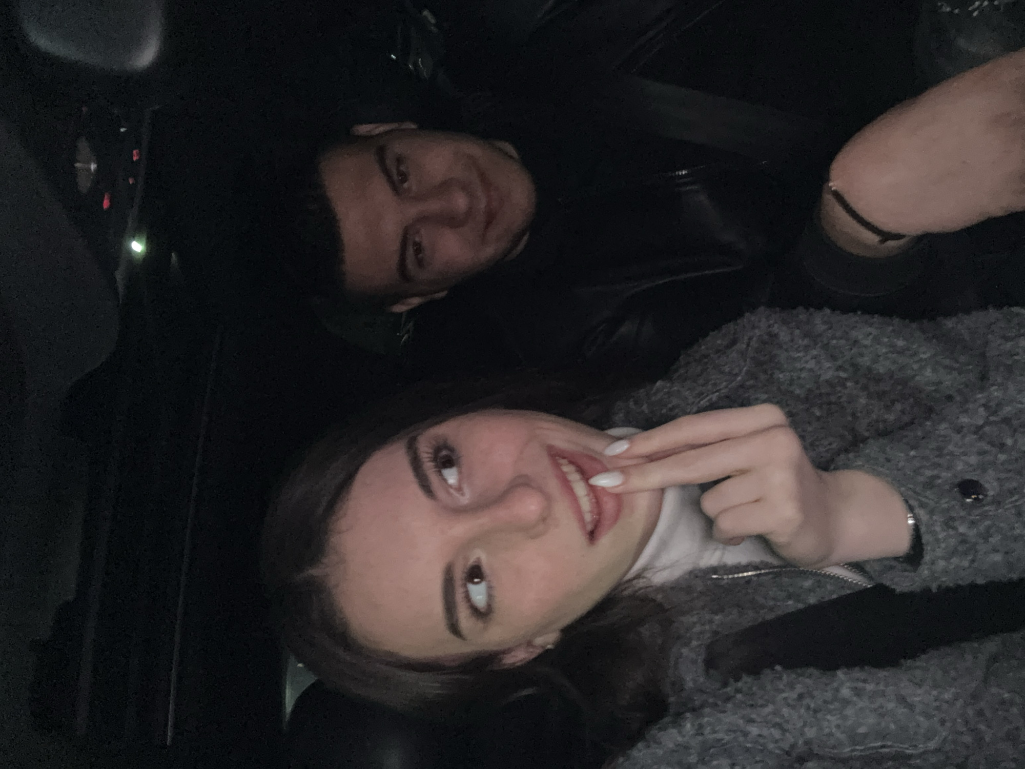 A low-angle, medium shot depicts a man and a woman inside a vehicle. The man is positioned in the upper-left corner of the frame, wearing a black leather jacket and a seatbelt, and he is smiling. The woman, occupying the lower portion of the frame, is wearing a gray textured coat over a white turtleneck shirt. She is smiling and holding her fingers near her teeth. A seatbelt is visible across her chest. The vehicle's interior is dark, with a glimpse of the dashboard visible in the upper-left corner. The specific location within Plovdiv, Bulgaria, is not discernable from the image.