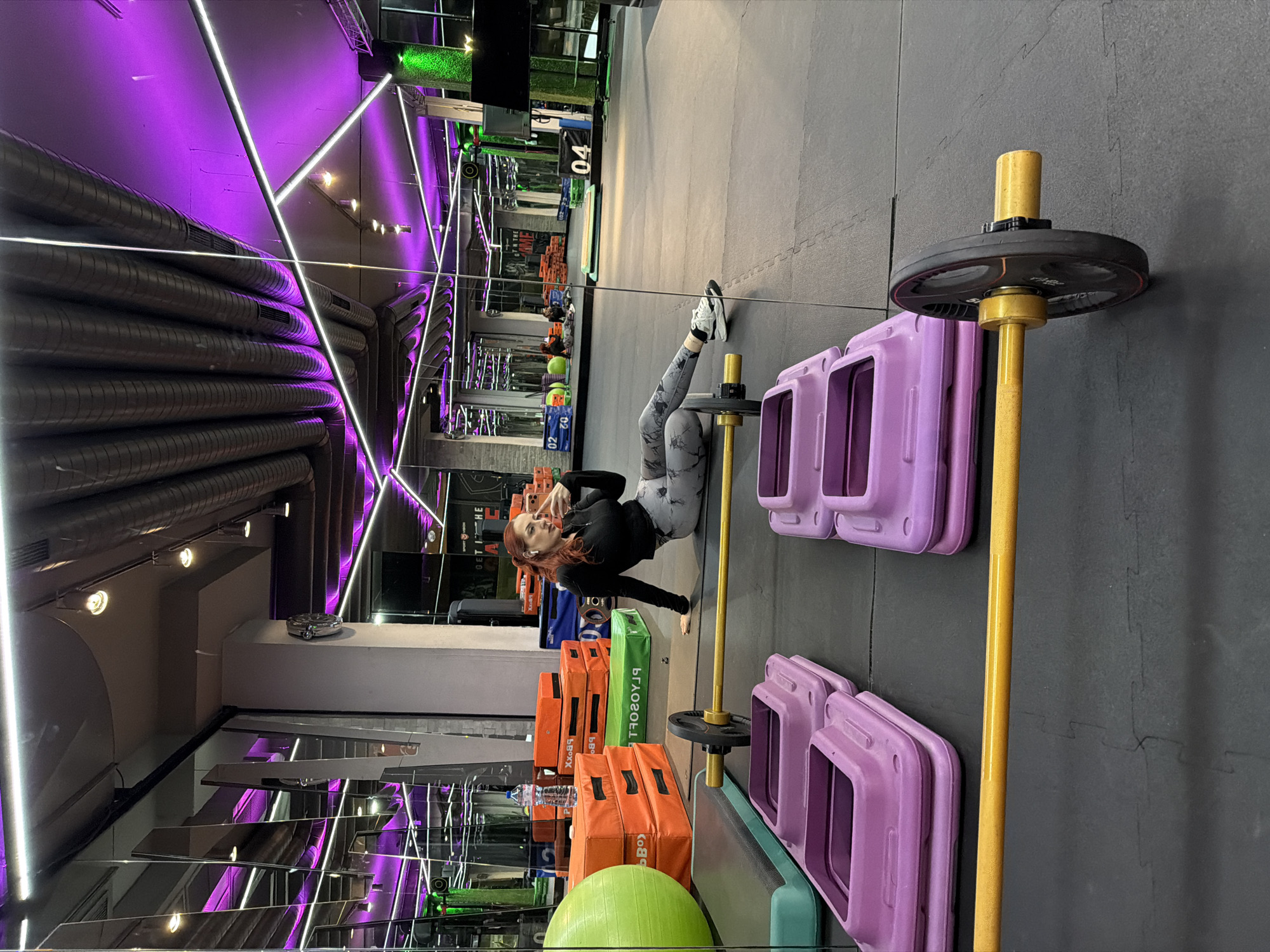 A woman is lying on her side in a modern fitness gym in Sofia, Bulgaria. She is facing towards the camera, wearing black workout attire and light gray leggings with white sneakers. Her red hair is loose around her shoulders. She is making a hand gesture with her right hand.
The gym features purple neon lighting, with reflective surfaces on the walls and ceiling. Exercise equipment, including violet step platforms and orange-colored storage boxes, is positioned nearby. A barbell stands upright. The floor is made of dark gray rubber mats.