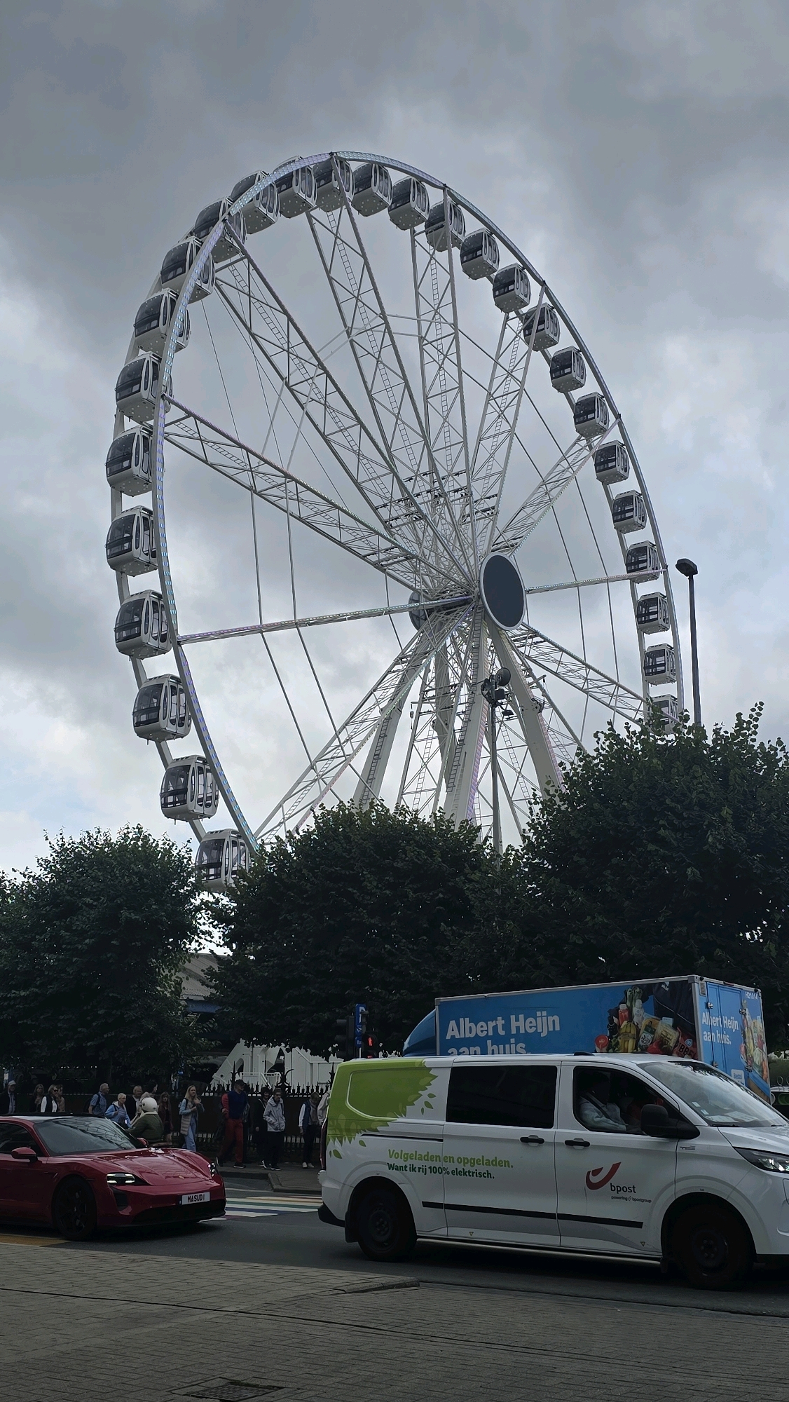 The image captures a scene in the city of Antwerp, Belgium, dominated by a large Ferris wheel. The Ferris wheel, with its intricate metal structure and numerous enclosed cabins, is positioned against a cloudy sky. In the foreground, two vehicles are visible on a paved road. On the left, a red sports car, identified by its license plate as "MASUDI", is parked. To the right, a white van with green foliage graphics and the text "Volgeladen en opgeladen. Want ik rij 100% elektrisch." and the Bpost logo is driving. Behind the van, a blue truck with the "Albert Heijn aan huis" logo is also present. A group of people are gathered on a sidewalk behind the red car, some appearing to be waiting or observing. Trees line the background, partially obscuring buildings. The overall atmosphere suggests an urban environment with a mix of leisure and commercial activities.