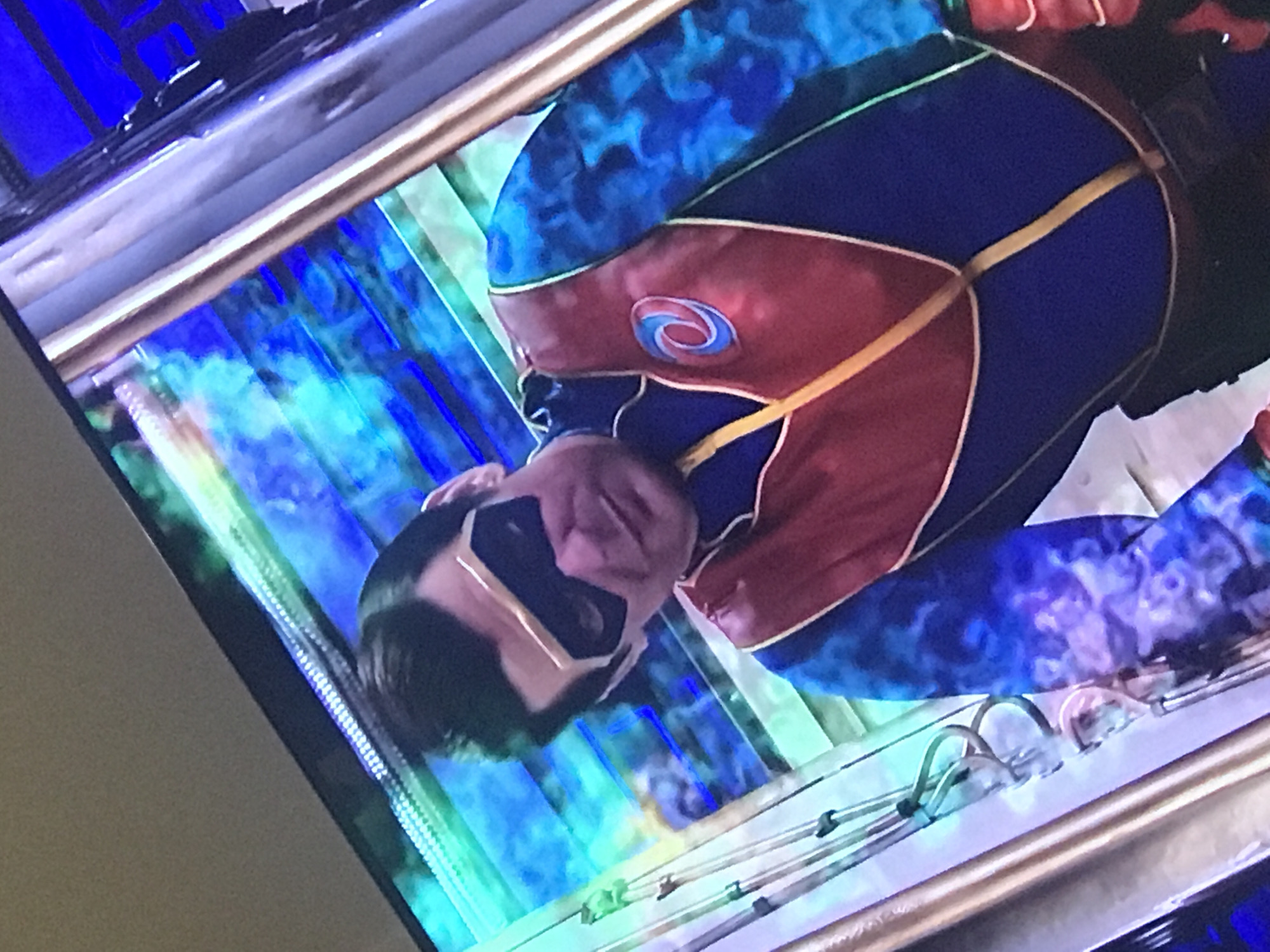 A screen shows a person in a superhero costume. The costume is primarily blue and red with gold detailing and a black mask. The backdrop appears to be a brightly lit, abstract design with blue and green tones. The screen is being photographed at an angle. The location is indeterminate and not necessarily Yerevan, Armenia.