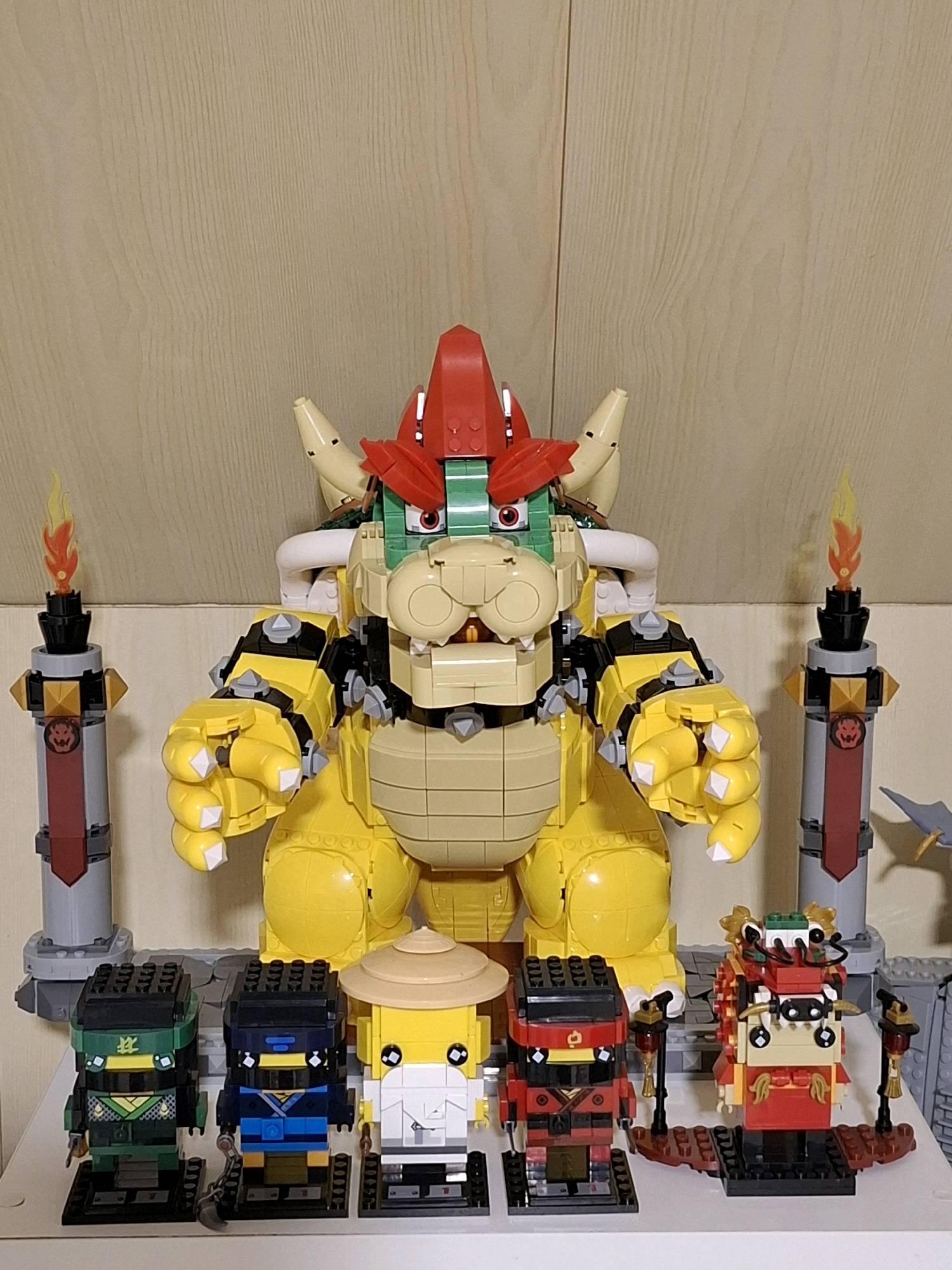 A large, yellow Lego Bowser figure dominates the center of the frame, standing on a grey Lego base. The figure is detailed with spikes, horns, and a red spiked shell. Flanking Bowser are two Lego torches with flame elements, one on each side. In front of Bowser, a row of smaller Lego figures are arranged. From left to right, there is a green and black ninja figure, a blue and black ninja figure, a figure in a straw hat, a red and black ninja figure, and a red and gold dragon-like figure. The background is a light brown wooden texture. The image appears to be taken indoors, on a shelf or table. There is no indication of the city of Tyumen, Russia, or any specific location.