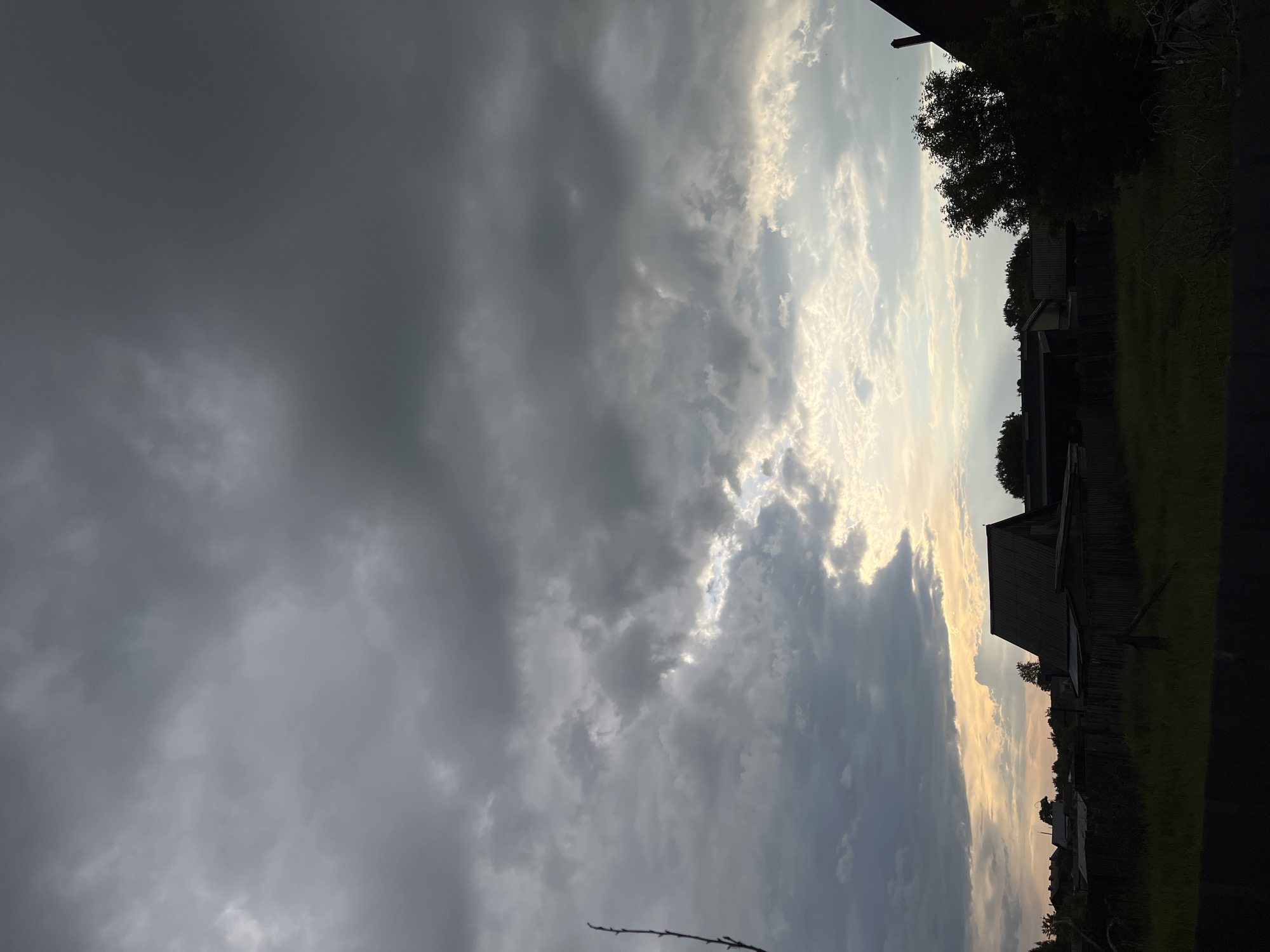 The image presents a late afternoon or early evening landscape in Novocheremshansk, Russia. The sky dominates the upper two-thirds of the frame, characterized by extensive, dark grey overcast cloud formations. Towards the lower middle and right, a significant break in the cloud cover reveals intense, yellowish-white light, indicative of a low sun or crepuscular rays breaking through.
The immediate foreground features a dark, indistinct horizontal barrier. Above this, a field or overgrown area of tall grass and sparse dry vegetation is rendered in dark silhouette. A horizontal wooden fence, also silhouetted, spans the midground. Beyond the fence, a series of rural structures, appearing as houses and outbuildings, are visible in silhouette against the brighter horizon. These buildings exhibit dark, varied rooflines, with a prominent, steeply pitched dark roof on a structure located on the right side of the frame. Interspersed among the buildings and along the horizon are silhouetted trees and bushes. A single, slender, bare branch extends vertically from the left edge of the image into the cloudy sky. There are no individuals, discernible actions, or interactions present within the scene.