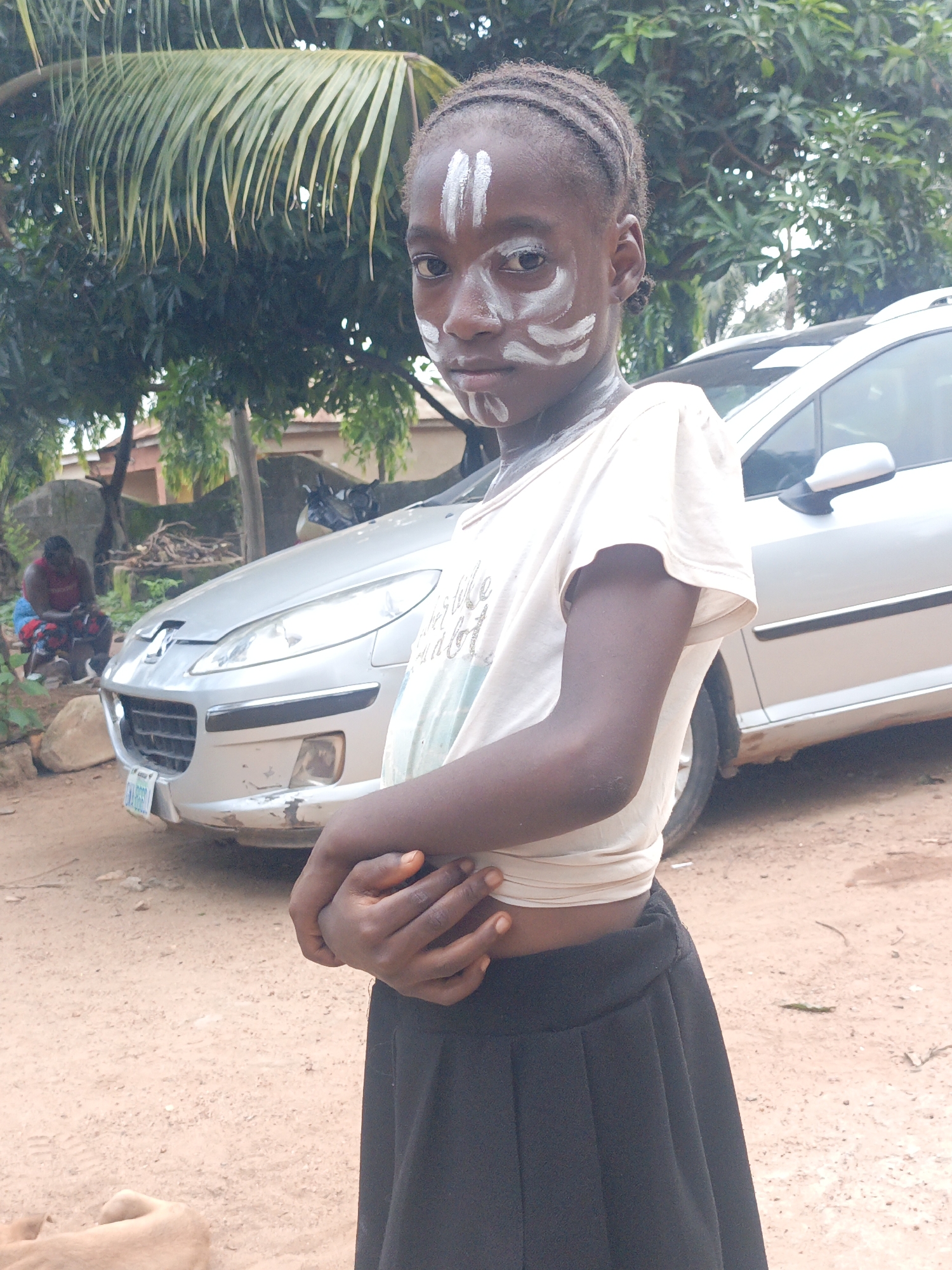 In Akwanga, Nigeria, a young girl with white face paint, stylized as lines and swirls, stands in a dusty outdoor area. She wears a beige, cropped t-shirt and a black skirt. Her arms are crossed in front of her stomach. Behind her, a silver sedan is parked. Further in the background, a woman in a red top and patterned skirt sits on the ground. Trees with large green leaves provide shade overhead, and buildings can be seen in the distance. The ground is composed of reddish-brown dirt and scattered debris.
