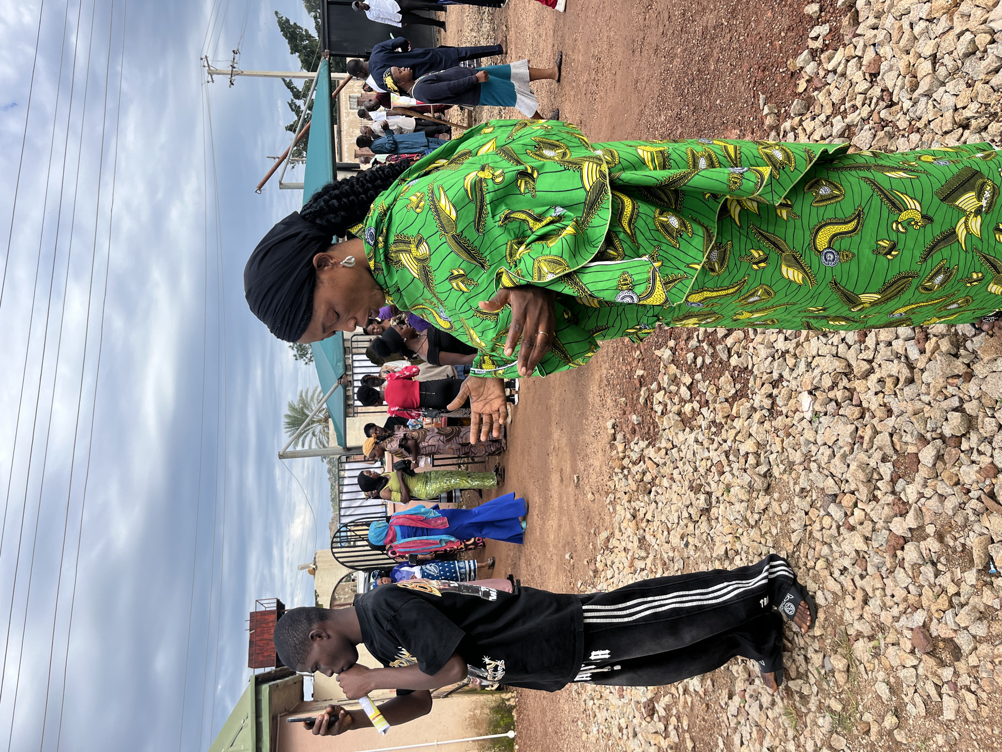 The image depicts an outdoor scene in Jos, Nigeria, characterized by a cloudy sky and a gravelly ground surface. In the foreground, a young man wearing a black t-shirt and black track pants with white stripes is holding a mobile phone to his ear, as if in a phone conversation. To his right, a woman in a vibrant green and yellow patterned traditional dress and a black headscarf is positioned with her arms in a prayer-like or gesture-holding pose. She is looking downwards. Behind them, a group of people are gathered, with some individuals wearing colorful attire, including a woman in a blue dress and a red headscarf, and another in a green top and patterned skirt. Several structures, including buildings with green roofs and a metal gate, are visible in the background. A utility pole with wires is also present. The overall atmosphere suggests a social gathering or event.