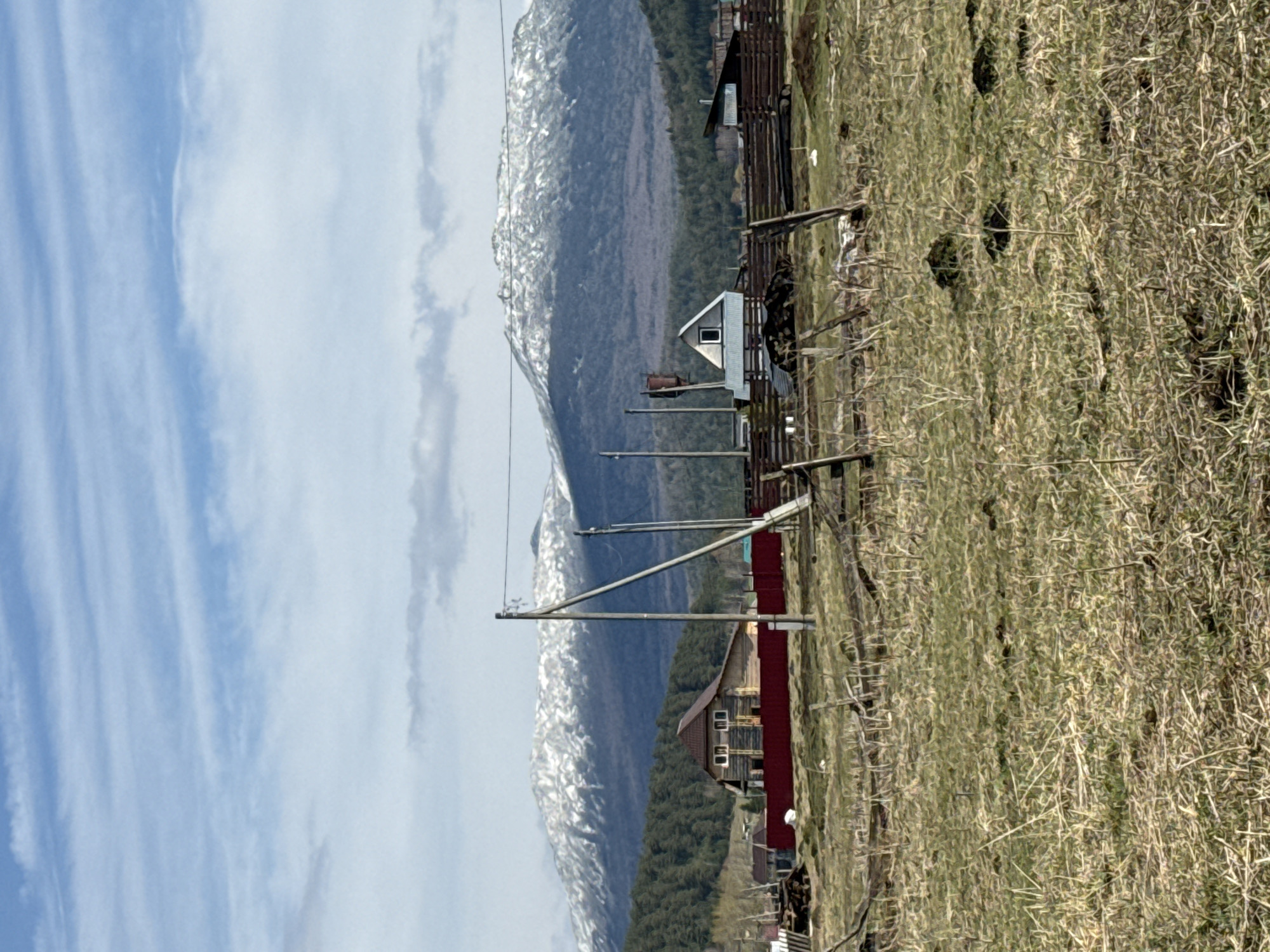 This image shows a rural scene in Tyulyuk, Russia. In the background are snow-capped mountains under a partly cloudy sky. In the midground, a wooden fence runs across a grassy field interspersed with dry brush. Several wooden structures, including houses and sheds with corrugated metal roofs, are visible. Power lines stretch across the foreground. The image has been rotated, making the scene appear sideways.