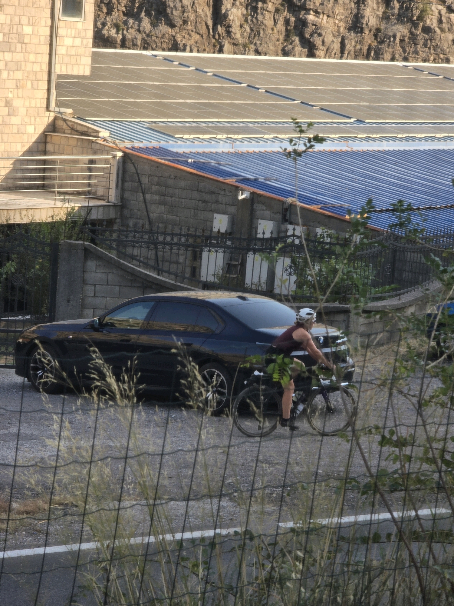 The image depicts a street scene in Podgorica, Montenegro, with a dark-colored sedan parked on the right side of the frame. A person wearing a white helmet and a maroon tank top and green shorts is riding a bicycle beside the parked car, appearing to be moving forward. The background features a building with a tiled roof and solar panels, a stone wall, a black metal fence, and a rocky hillside. Dry grasses and foliage are visible in the foreground, partially obscuring the view of the car and cyclist. The lighting suggests it is daytime, possibly late afternoon.