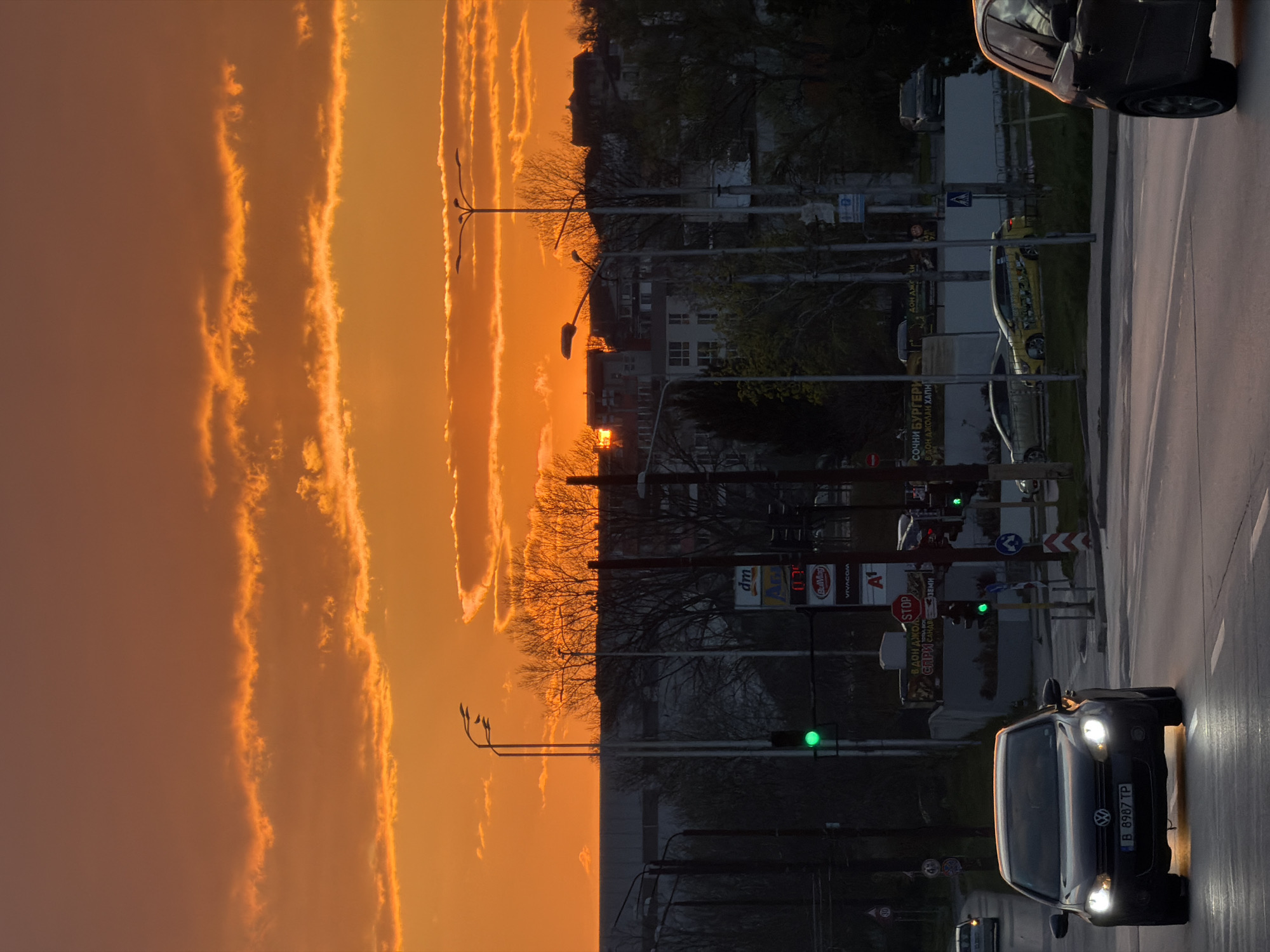 The image captures a street scene in Varna, Bulgaria at sunset. The sky is a vibrant orange with streaks of clouds reflecting the light. On the right side of the image is a street with cars, traffic lights, and commercial signs. A dark-colored car is visible in the foreground with its headlights on, and a license plate that reads "B 8987 TP." Above the car, there are signs for "dm", "BulMag", "VIVACOM" and "A1", next to traffic signs "STOP". There is a traffic light with a green light illuminated. The scene is bisected by the boundary between the sky and the urban environment, with trees and buildings forming a silhouette against the sunset. Streetlights are interspersed between the sky and the built environment. The overall impression is of a city street bathed in the warm hues of a late sunset.