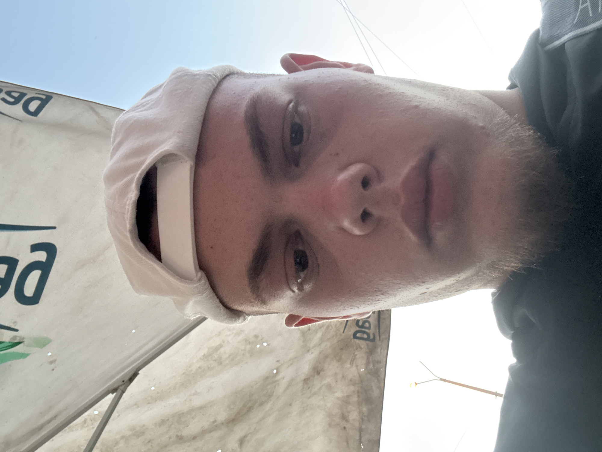 A close-up, low-angle self-portrait of a young man wearing a light-colored baseball cap. The man has fair skin, light-brown hair, and a short beard. He is looking directly at the camera. The background includes a white tent or canopy with the letters "ga" visible in blue, suggesting it is a logo or part of a word. In the far background, a street lamp and light-colored sky are visible. It is difficult to specify the exact location in Burgas, Bulgaria, without additional details or landmarks.