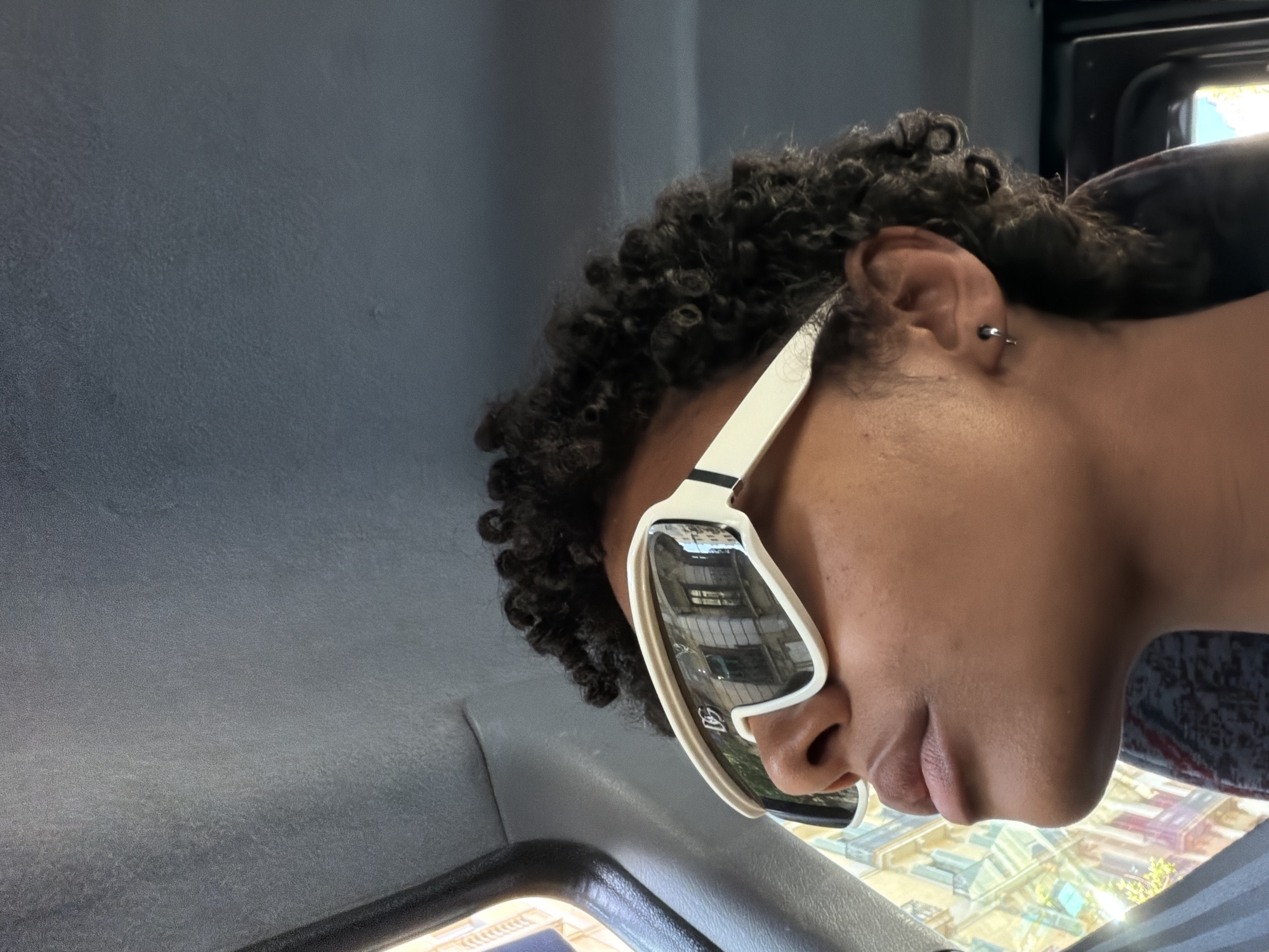 A close-up, angled view of a person in a vehicle, possibly a bus or taxi, is depicted. The individual has curly black hair, dark skin, and is wearing white-framed sunglasses that reflect a cityscape. A small, silver hoop earring is visible in the person's ear. The vehicle's interior, with a gray ceiling and window frame, is visible. In the reflection of the sunglasses, buildings are discernible. The scene is in Barcelona, Spain, indicated by the architectural style reflected in the sunglasses.