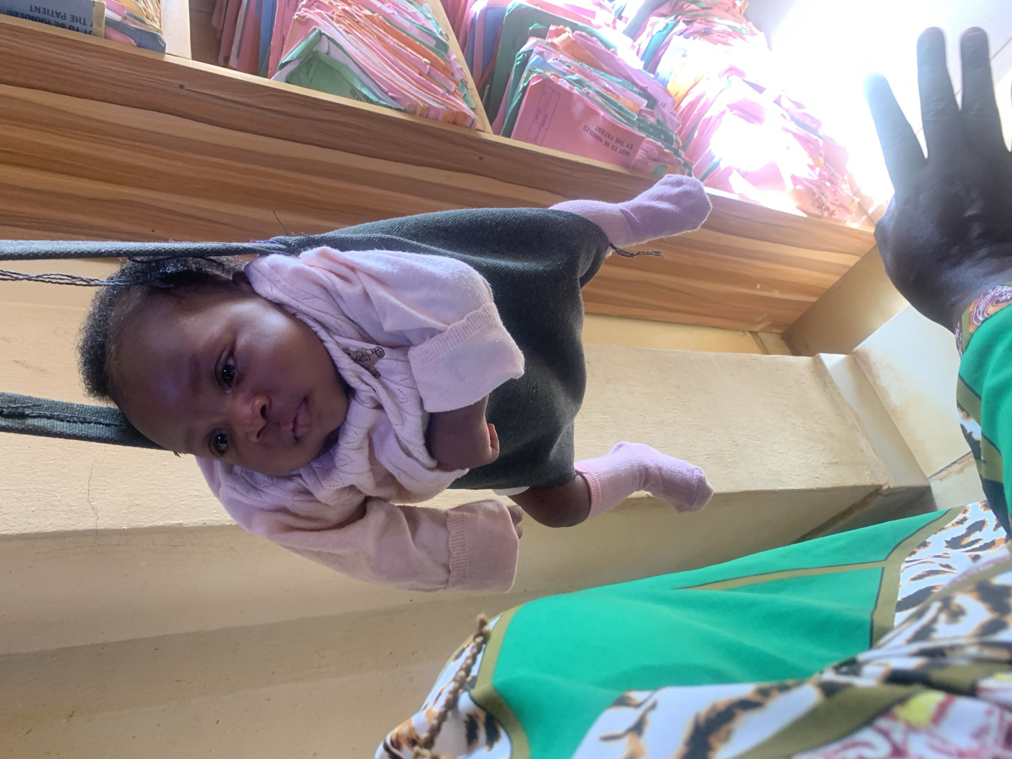 The image captures a unique low-angle perspective, looking upwards at a young baby suspended in a dark grey fabric carrier or wrap. The baby, with dark hair and eyes, is dressed in a light pink long-sleeved top and pink socks, and gazes calmly towards the viewer. Its head is positioned towards the bottom left of the frame, and its body extends upwards.
To the right, a dark-skinned adult's arm and hand are visible, seemingly supporting or interacting with the baby. The adult is wearing a vibrant green garment with a colorful, patterned trim.
In the background, a wooden shelf or ledge is prominently featured, laden with numerous stacked files and folders. These files, appearing to be administrative or medical records, are primarily pink and green, with other assorted colors. Visible text on some of the pink folders clearly states "NOT TO BE HANDLED BY THE PATIENT" and "THE PATIENT," strongly suggesting a medical or record-keeping environment, such as a clinic or hospital in Jos, Nigeria.
Bright light illuminates the scene from above the shelf, creating a diffused glow. The wall below the baby and behind the adult's arm is a light, neutral tone. The image conveys a tender moment of interaction, with the baby's presence in an unexpected administrative setting.