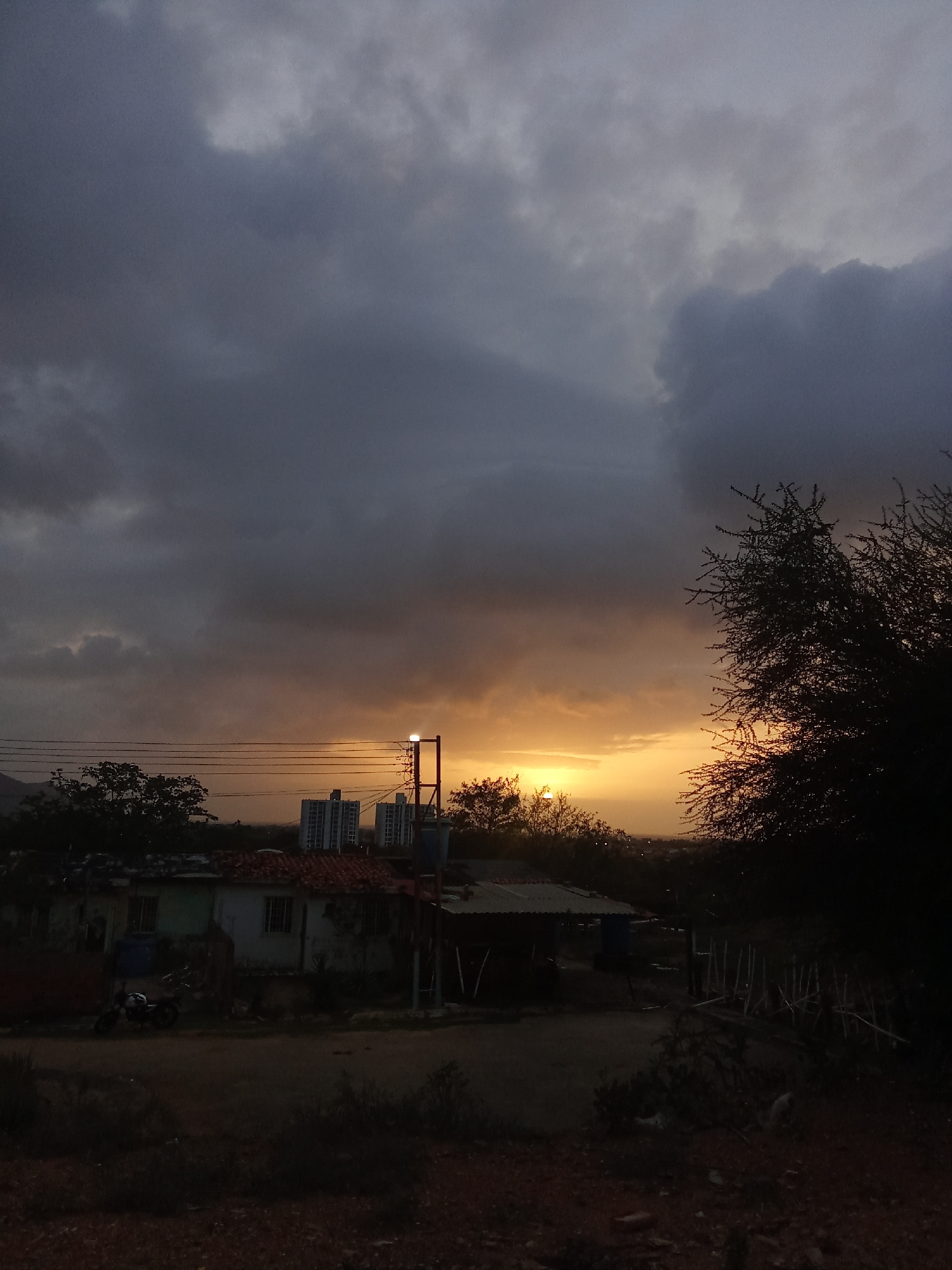 The image captures a twilight scene in the city of Juan Griego, Venezuela. A dramatic sky filled with dark, heavy clouds dominates the upper portion of the frame, with a sliver of warm, orange sunset light breaking through near the horizon. In the mid-ground, a collection of buildings is visible. Two tall, white, modern structures stand out against the more rustic architecture of the foreground. A single-story building with a red-tiled roof and light-colored walls is situated closer to the viewer. A streetlight with a glowing bulb stands prominently on a pole, casting a soft light. Power lines crisscross the scene horizontally. To the right, the silhouette of a thorny tree is starkly outlined against the colorful sky. The foreground consists of a dirt or gravel path, with sparse vegetation and debris scattered on the ground. A motorcycle is parked on the left side of the path, near the single-story building. The overall atmosphere is subdued, indicative of the transition from day to night, likely just before or during a rain shower given the cloud formation.