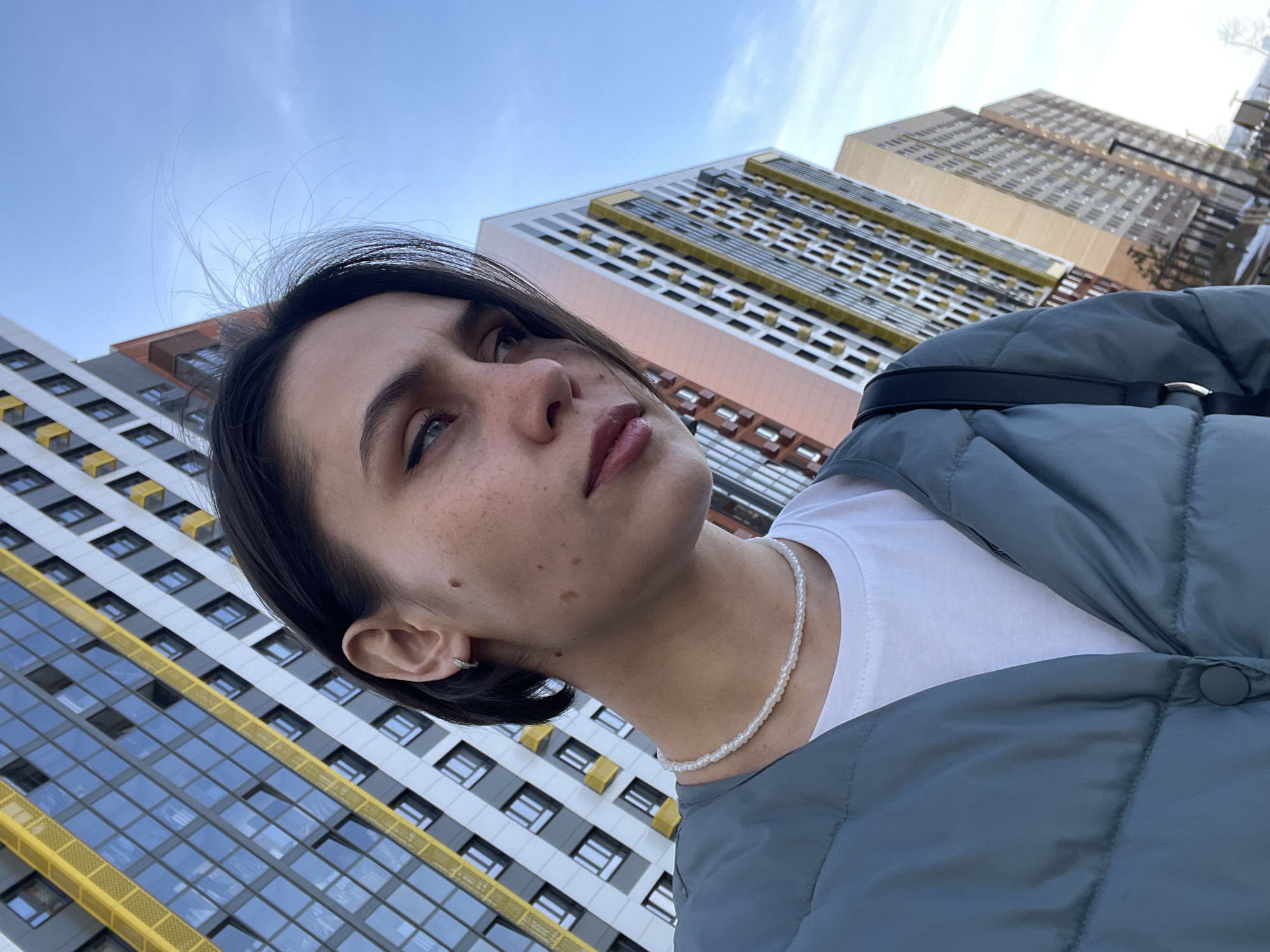 A low-angle medium shot captures a woman against a backdrop of modern high-rise buildings. The woman, positioned on the left, has short dark hair, fair skin with freckles, and is wearing a white t-shirt, a pearl necklace, and a blue-gray puffer jacket. The buildings in the background have a grid-like pattern with multiple windows. The sky above is light blue with faint clouds. The location is the city of Izhevsk, Russia.