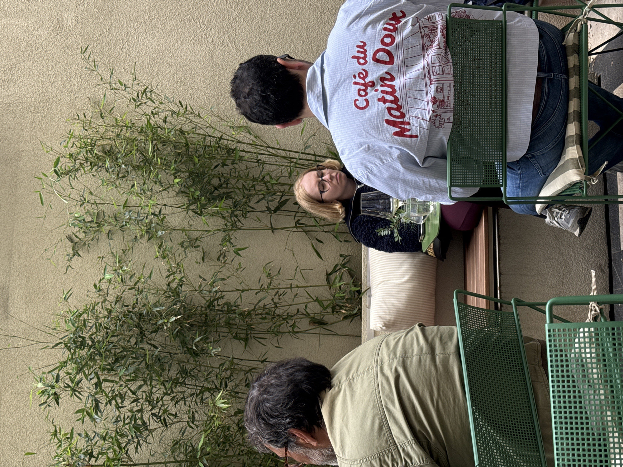 Three individuals are seated outdoors at a cafe in Belgrade, Serbia. A man in a light blue shirt with "Café du Matin Doux" printed in red on the back sits facing away from the camera. A woman with blonde hair and glasses sits beside him, looking towards the camera. An older man with gray hair and glasses sits facing the first man. They are all seated in green metal chairs. A green leafy plant is positioned behind them, partially obscuring a light-colored wall. There is a table with drinks.