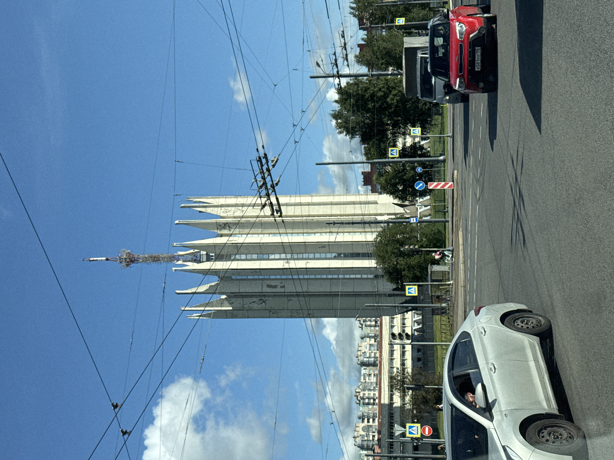 The image depicts an urban street scene in Saint Petersburg, Russia, featuring a prominent, tall, angular building resembling a television tower or communication tower, topped with an antenna. The tower is white and has a distinct, pointed architectural style. In the foreground, a multi-lane asphalt road is visible, with traffic lights and overhead wires for trolleybuses crisscrossing the sky. Several vehicles are present, including a light-colored sedan in the left lane, a red compact SUV in the right lane, and a gray van behind it. Pedestrians are seen near crosswalks, and a person on a bicycle is visible in the middle ground. The sky is blue with scattered white clouds. In the background, a row of apartment buildings with varied architectural styles lines the horizon. The overall scene suggests a busy urban intersection on a clear day.