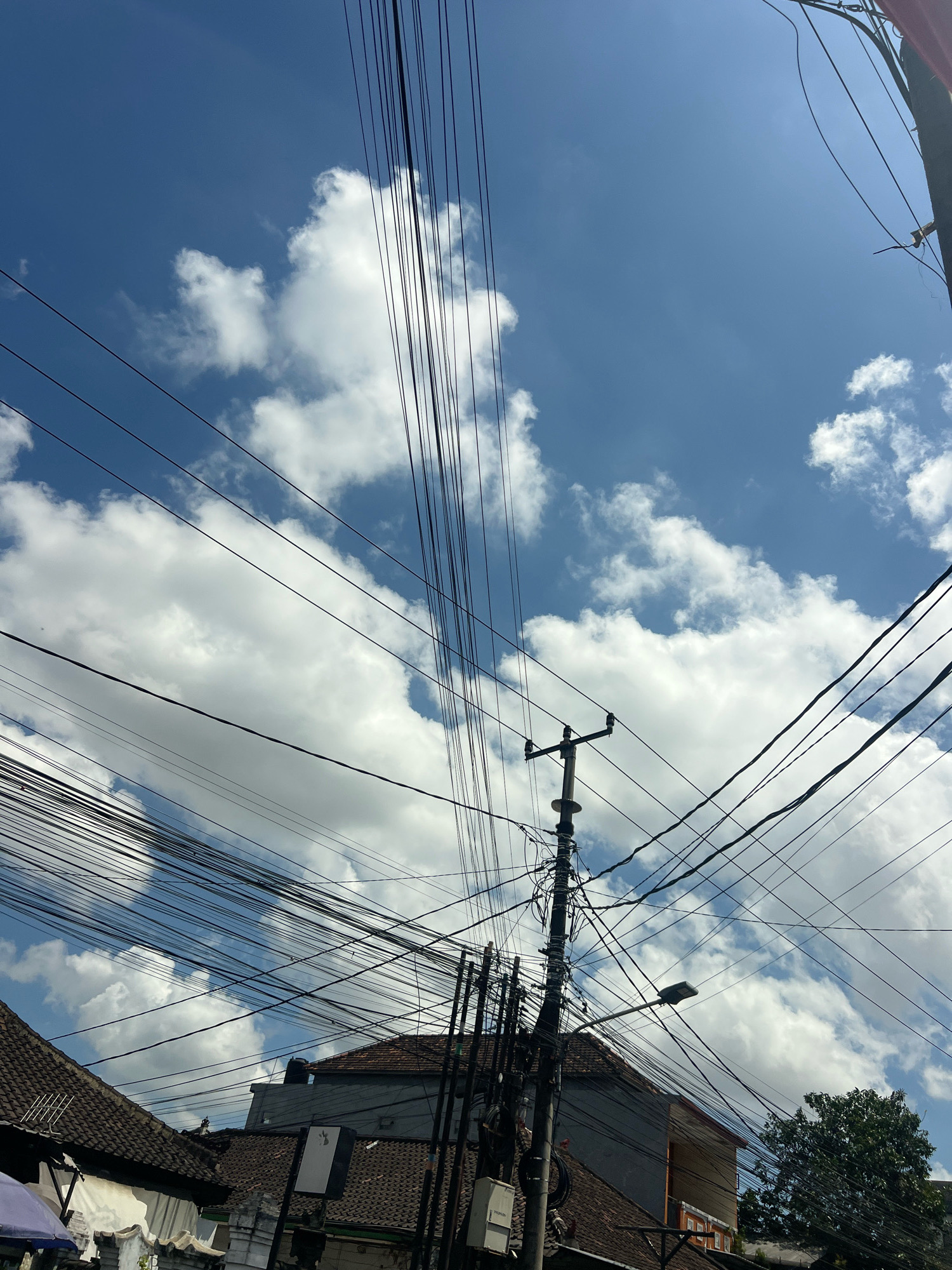 The image captures a view looking upwards at a tangle of electrical wires against a bright blue sky with fluffy white clouds. In the lower portion of the image, rooftops of buildings with dark, textured tiles are visible, along with a utility pole adorned with numerous wires and a streetlamp. A glimpse of a green tree can be seen on the right side. This scene is characteristic of the densely wired infrastructure often found in urban areas like Denpasar, Indonesia.