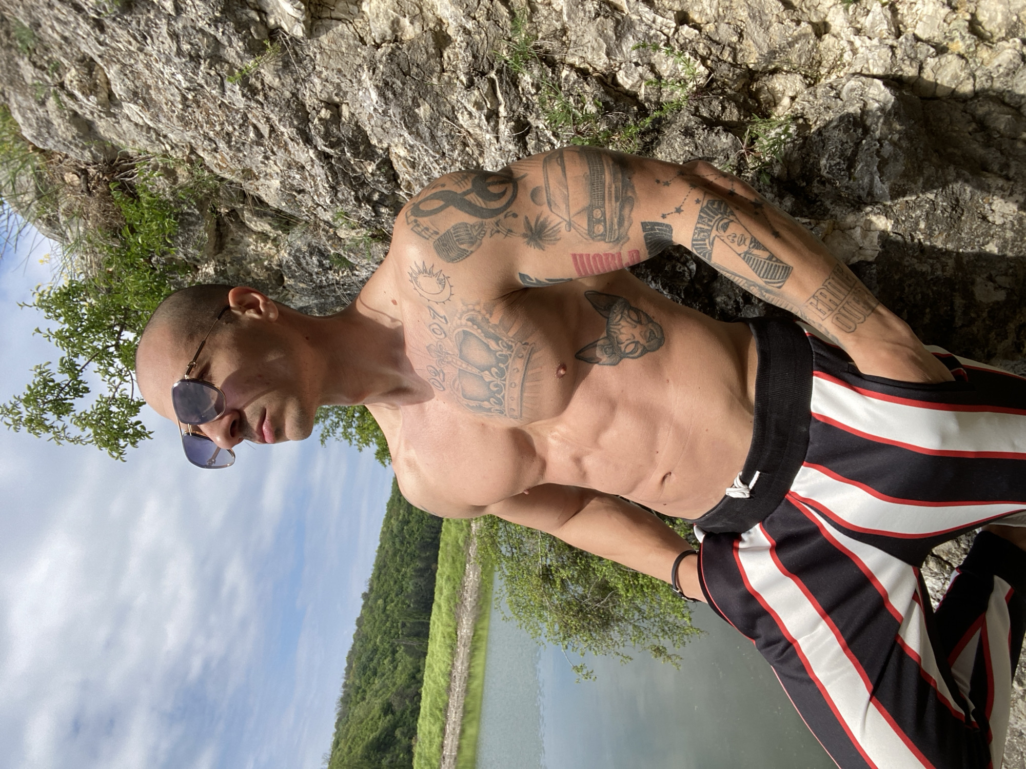 A man is posing outdoors on a rock formation, revealing his tattooed torso and upper body. He has a shaved head and is wearing sunglasses and striped athletic pants. The backdrop includes a body of water, greenery, and a rocky cliff face. The location appears to be in a natural environment, potentially near a lake or river in Pleven, Bulgaria.