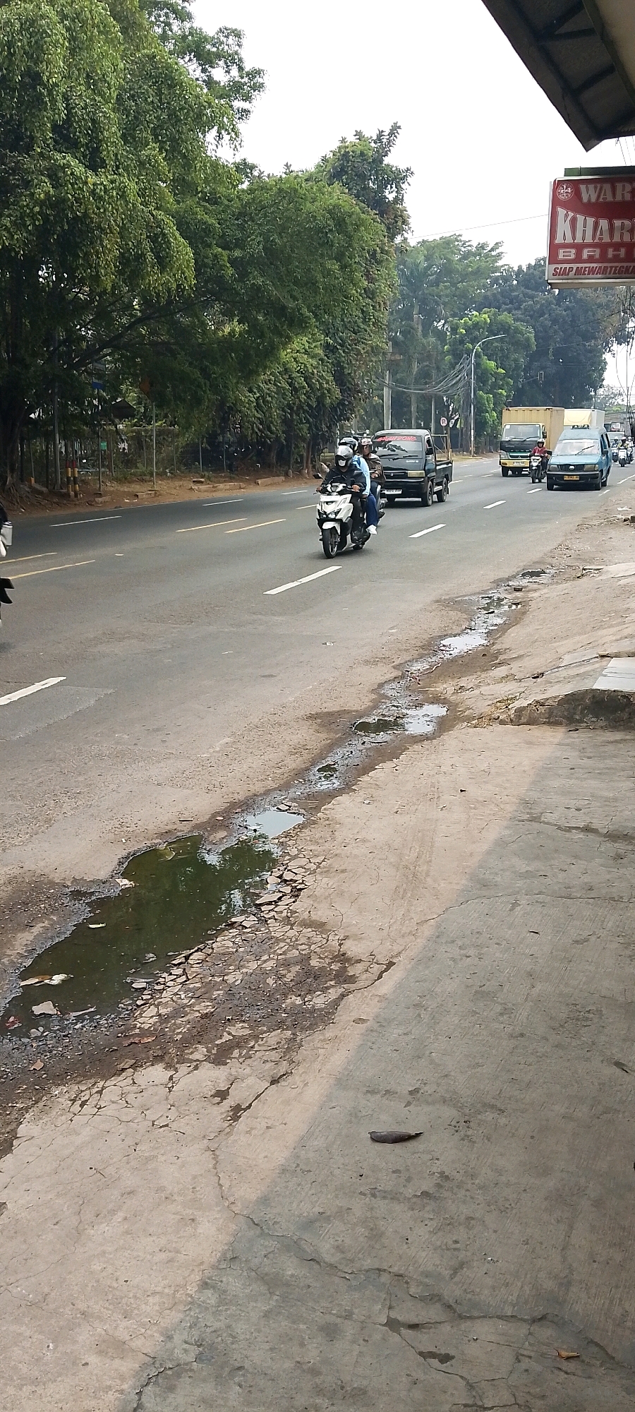 A side view of a road in Depok, Indonesia shows a few vehicles. In the foreground, on the right side of the image, there is a concrete sidewalk with several cracks and puddles of dark water, the largest of which is on the left side of the sidewalk. A single brown leaf rests on the sidewalk. The road itself has white lane dividers and some patchy areas with exposed gravel. A white motorcycle with two riders wearing helmets is in motion on the road, traveling away from the camera. Behind the motorcycle, a dark colored pickup truck is also moving away from the camera. Further in the background, a yellow truck and a blue car are visible, along with more trees and utility poles. A red and white sign is visible on the right side of the image, partially obscured by the roof of a building. The sky is overcast.