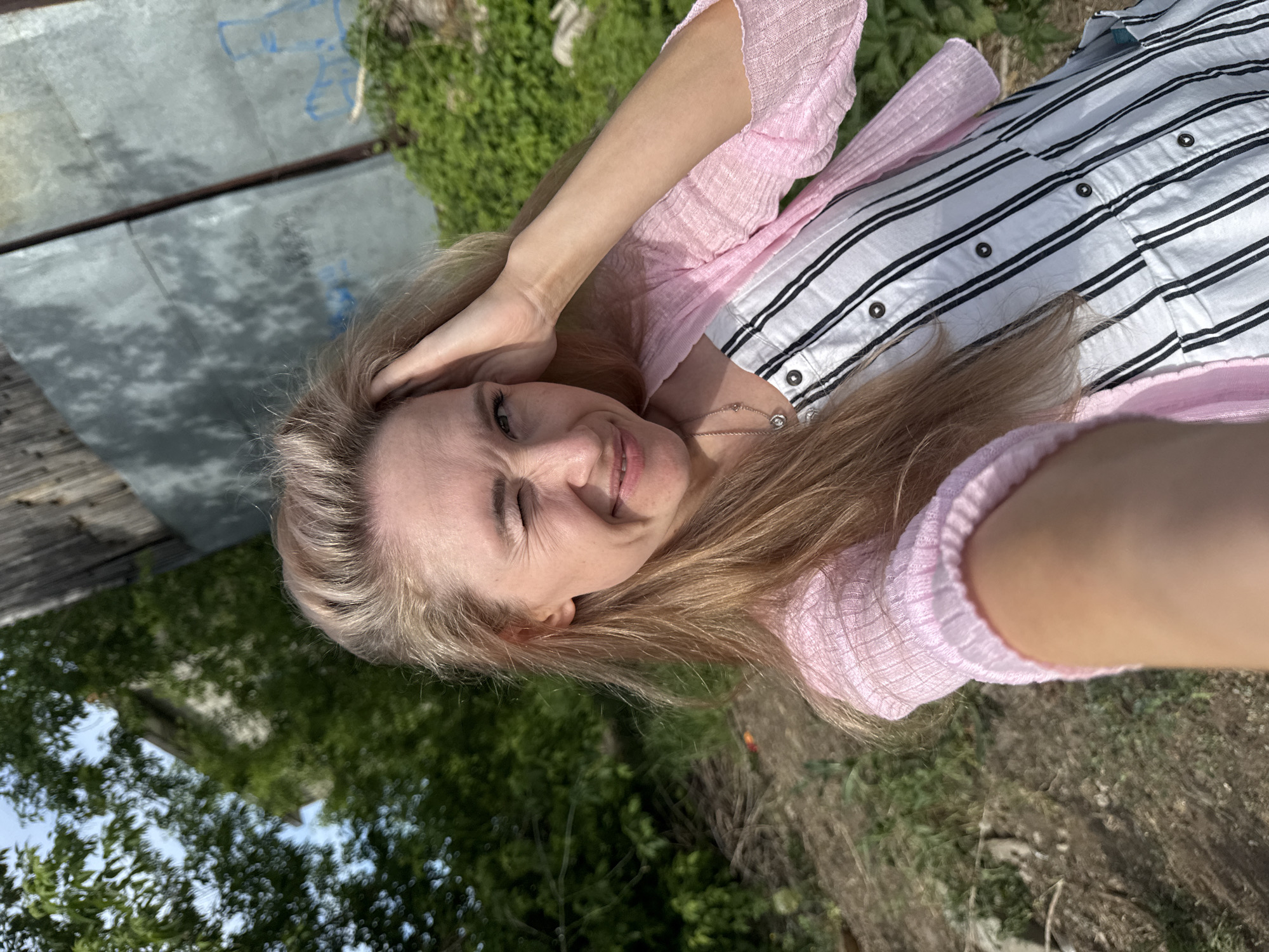 A fair-skinned woman with long blonde hair poses outdoors, winking and smiling while touching her hair. She wears a pink cardigan over a white, vertically striped button-down shirt. A thin necklace is visible around her neck. The background includes greenery and a structure with weathered wood and a gray, metal wall, possibly in the city of Chaikovsky, Russia. The ground appears to be dirt and grass. The image suggests a sunny, natural setting.