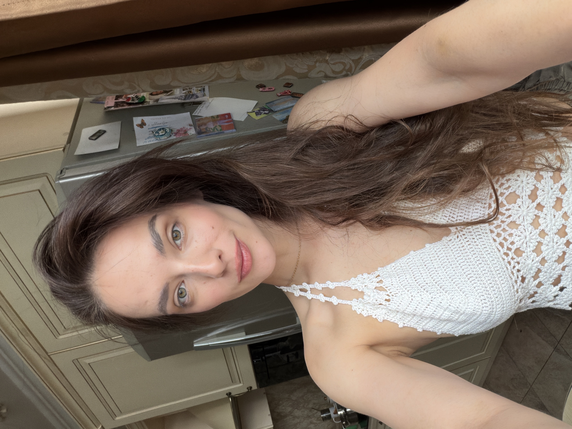A young woman with long, dark brown hair and light green eyes is taking a selfie in what appears to be a kitchen. She is wearing a white crocheted halter top. Behind her, a stainless steel refrigerator is visible, adorned with several pieces of paper and magnets. To her left are kitchen cabinets and a countertop with some items on it. To her right, a brown curtain hangs. The lighting suggests it is daytime.