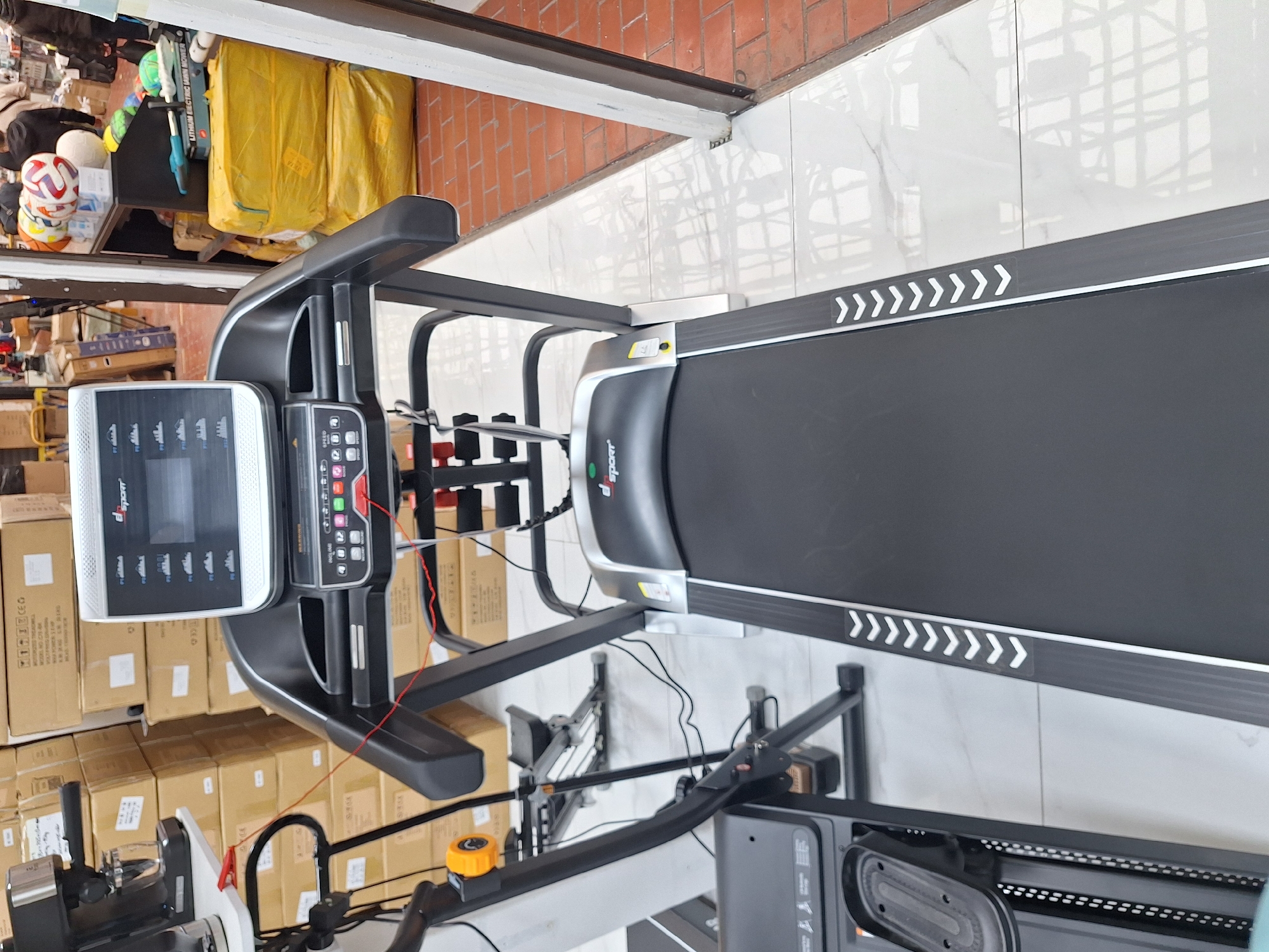 The image shows a treadmill and a rowing machine. The treadmill is black with a gray frame and a display panel. It has white arrows along the edge of the tread. The rowing machine is gray and black and has a seat and handlebars. The background is a cluttered space with boxes, bags, and other items. The scene is likely indoors in a retail or storage environment. There is no indication of being in Moscow, Russia.