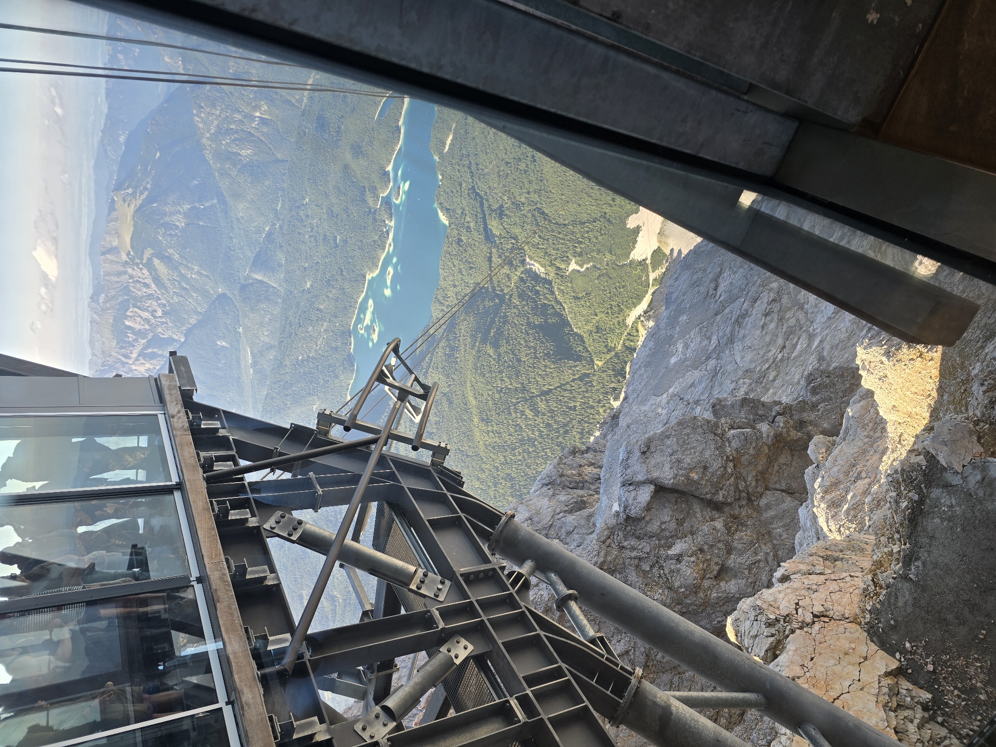 This is a high-angle, full shot of a modern building structure with a panoramic view of a mountainous landscape. The building, made of metal beams and glass, extends from a rocky mountain peak. Through the glass, people are visible standing inside. The landscape features a turquoise lake surrounded by lush green forests and majestic mountains, possibly the Eibsee in Garmisch-Partenkirchen, Germany, viewed from the Zugspitze. The image captures the contrast between the man-made structure and the natural beauty of the Alps.