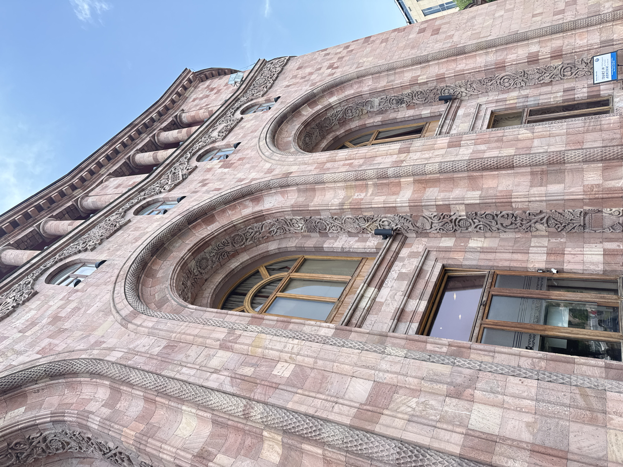 The image shows the facade of a building in Yerevan, Armenia. The building has an ornate architectural style, featuring pinkish-brown stone blocks, arched windows, and detailed carvings. The sky is visible in the background, indicating it is daytime. A sign with the words "Tours in Armenia" is visible on the building.