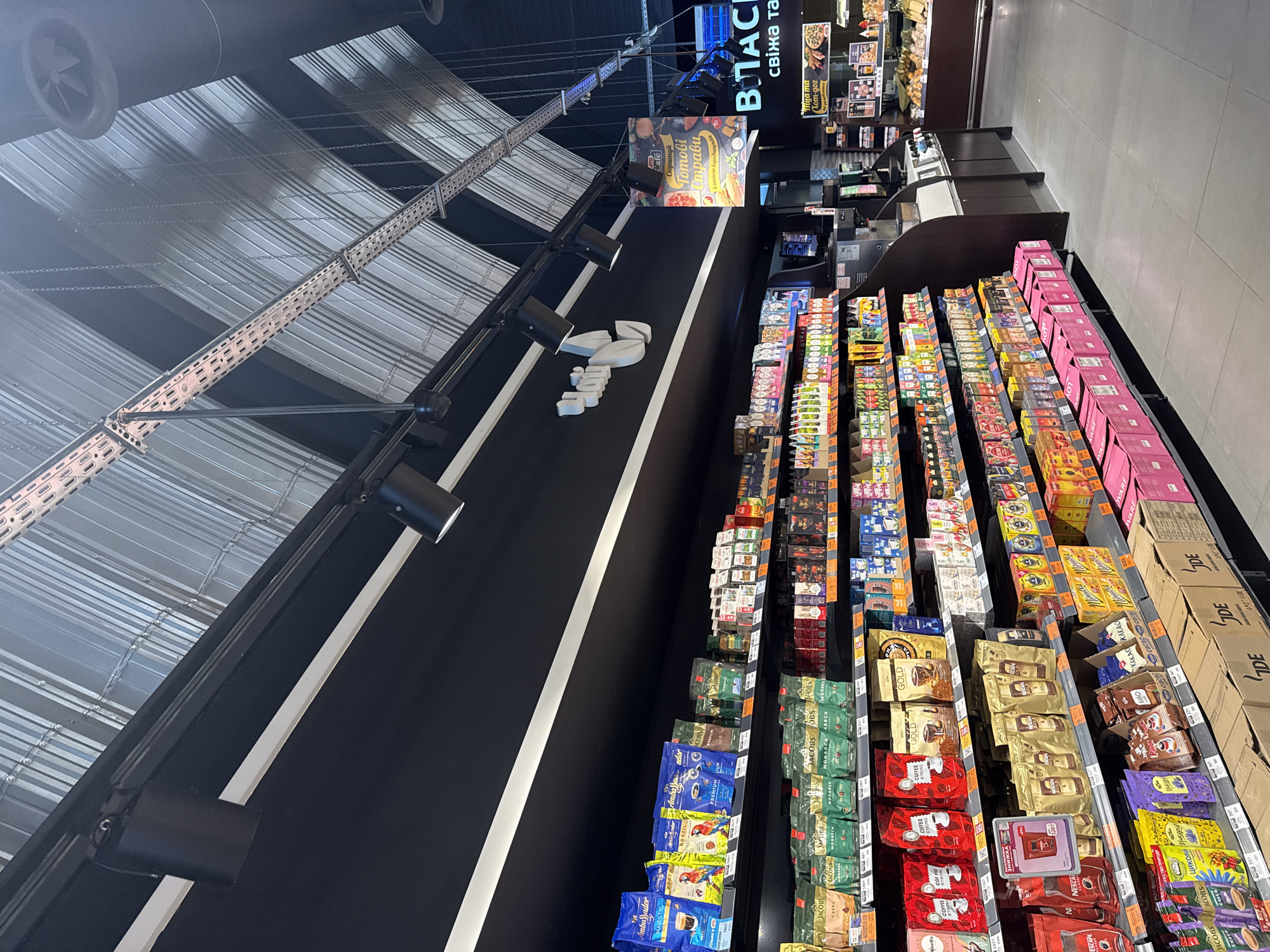 The image shows a well-lit interior of a supermarket in Dnipro, Ukraine. The camera is positioned at a slightly upward angle, capturing a section of the store's aisles and ceiling. The ceiling features a modern, curved design with metal struts and track lighting. Shelves are stocked with various food products, primarily coffee and tea brands, displayed in colorful packaging. Several rows of shelves are visible, filled with bags of coffee, boxes of tea, and other grocery items. Some shelves have price tags or labels. In the background, there's a checkout counter area with electronic devices, and a display of fresh produce like fruits and vegetables. The overall impression is of a clean, organized, and contemporary retail environment. There are no people visible in the image.